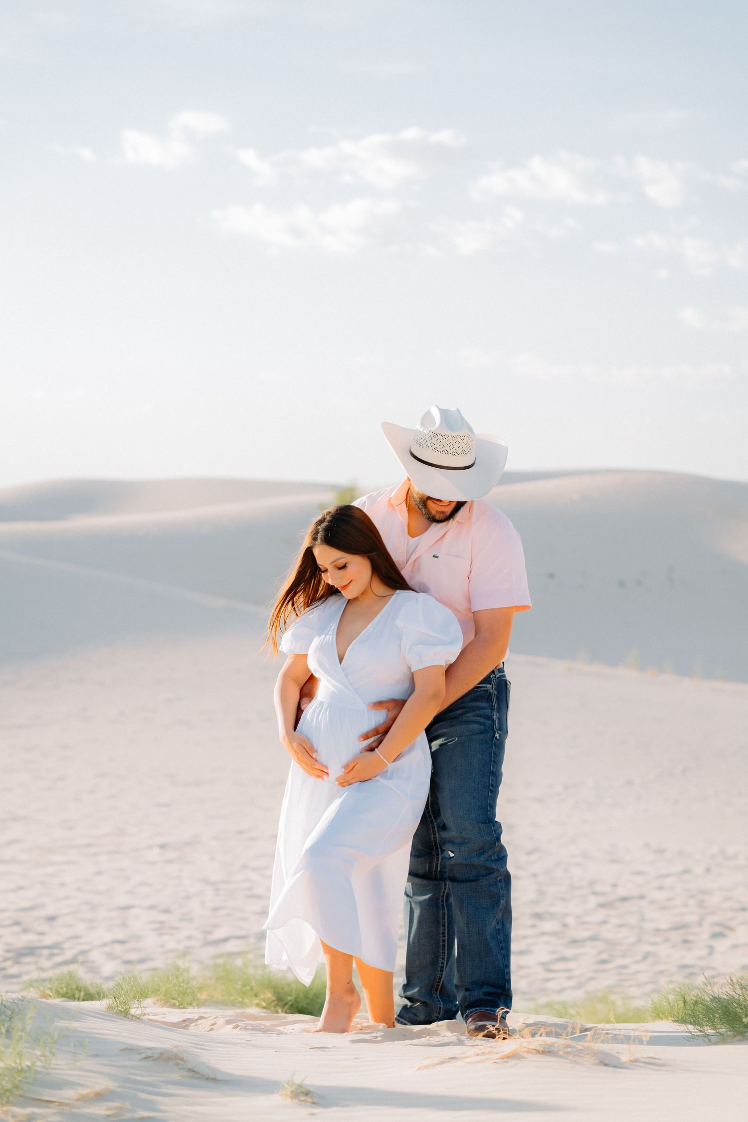 A couple in light summer clothing sharing an intimate moment on sand dunes at sunset.