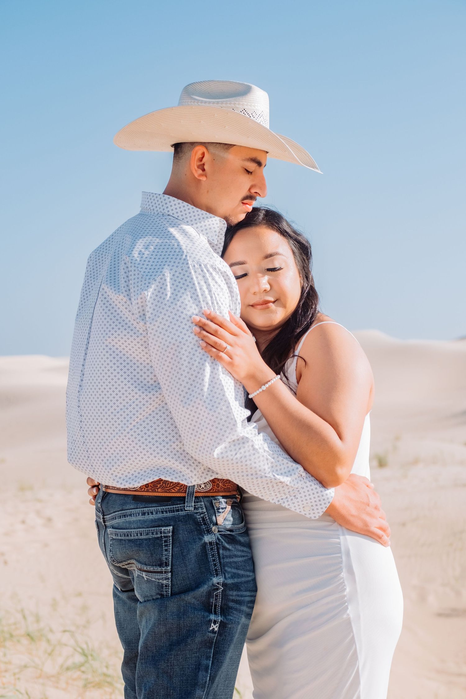 Couple in western attire sharing intimate moment on sandy dunes.