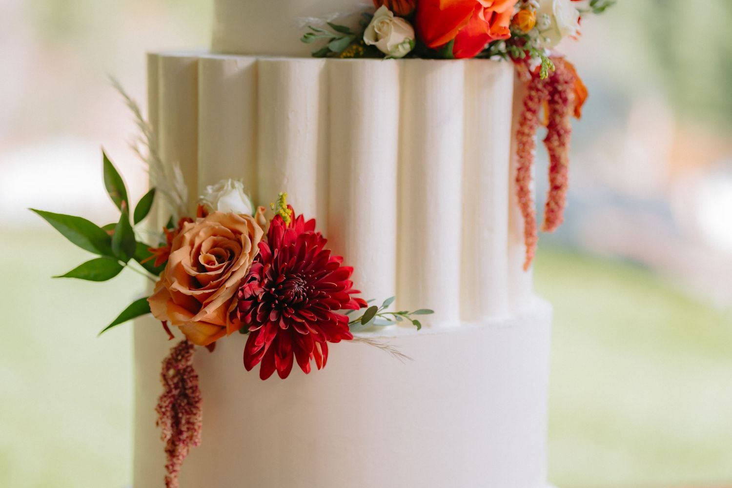 Close-up of white wedding cake tier decorated with fresh coral roses, dahlias, and cascading greenery.