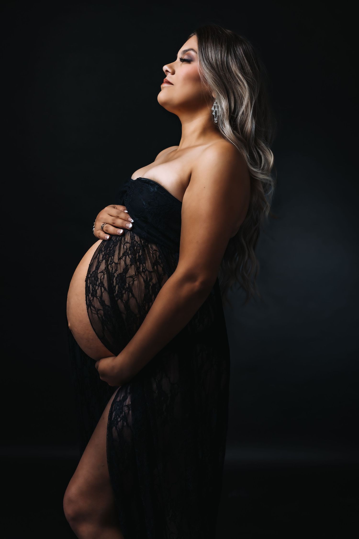 Artistic maternity photoshoot in black lace against dark background with dramatic lighting.