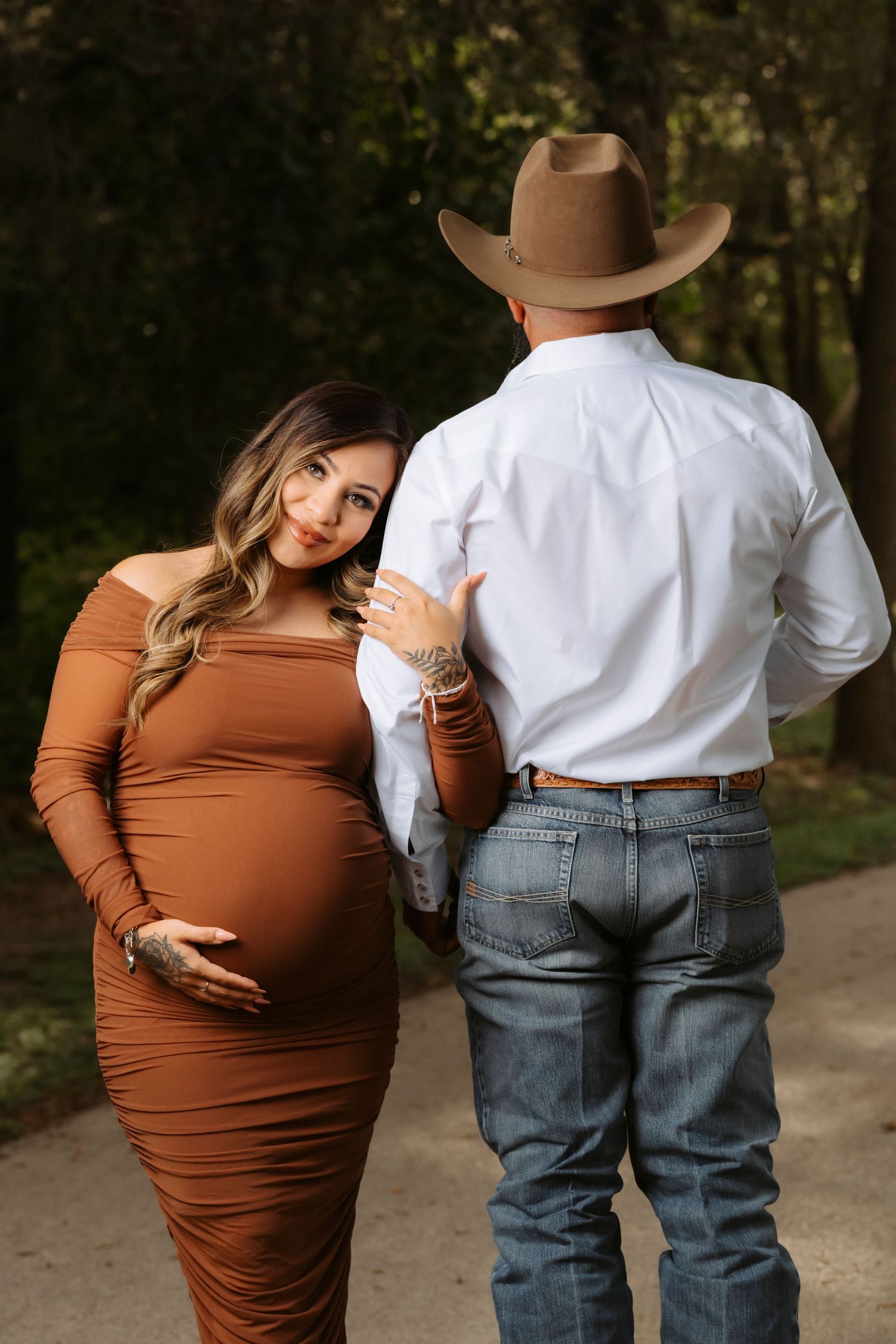 A couple in western attire poses outdoors, with a brown dress and cowboy hat creating a rustic maternity photo.