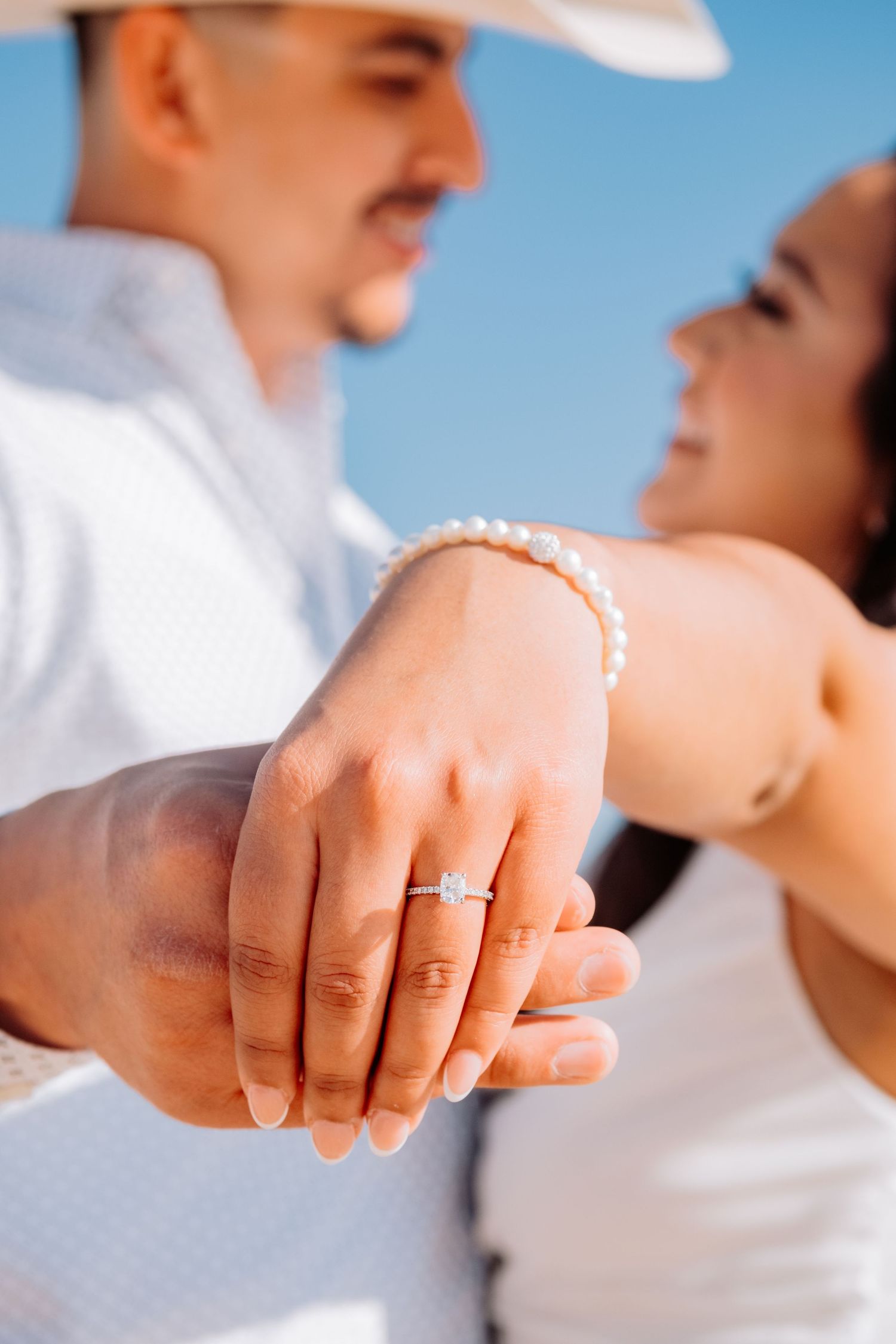 Close up of engagement ring and pearl bracelet with couple embracing in background.