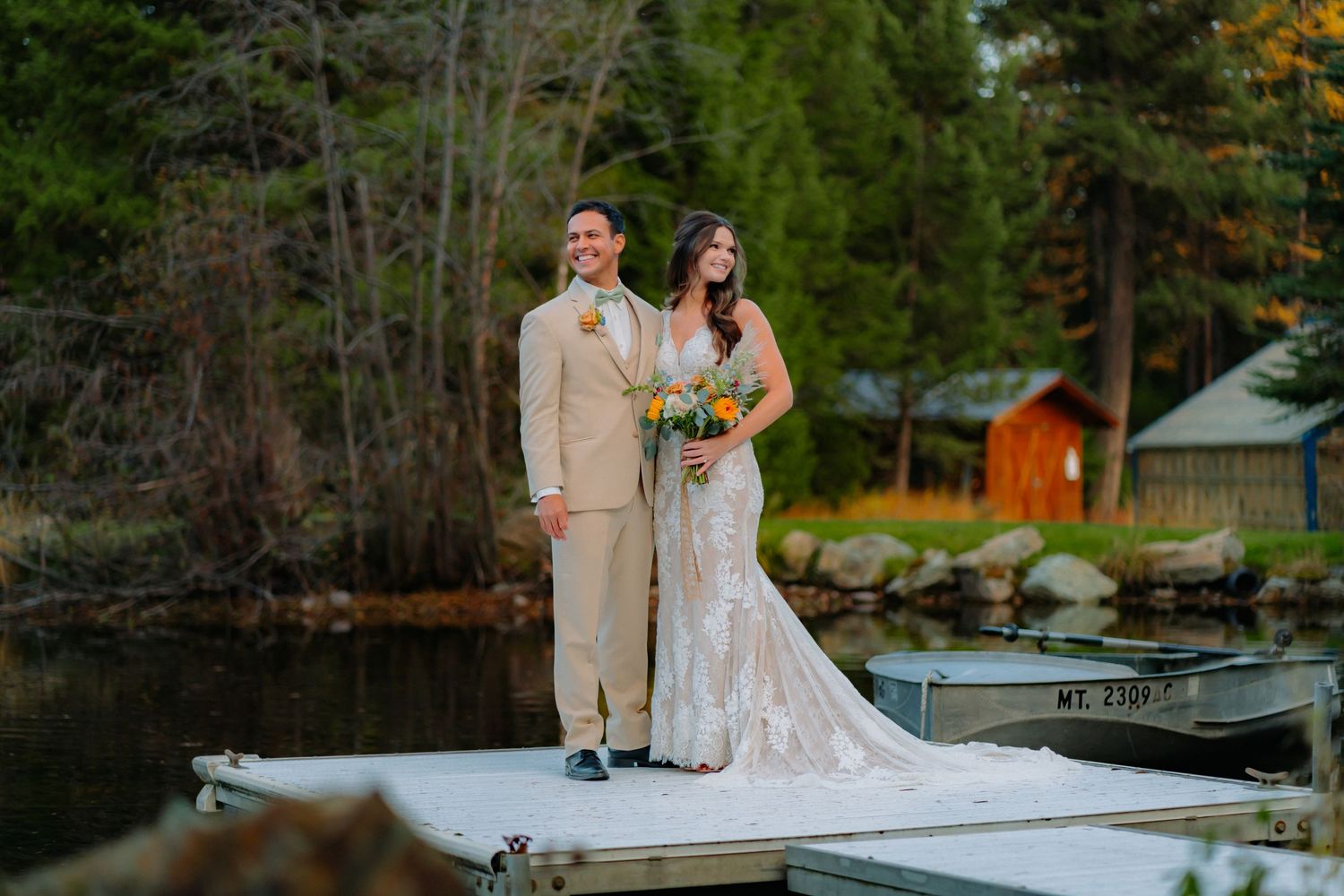Newlyweds pose together on wooden dock with rustic cabin and autumn trees in background.