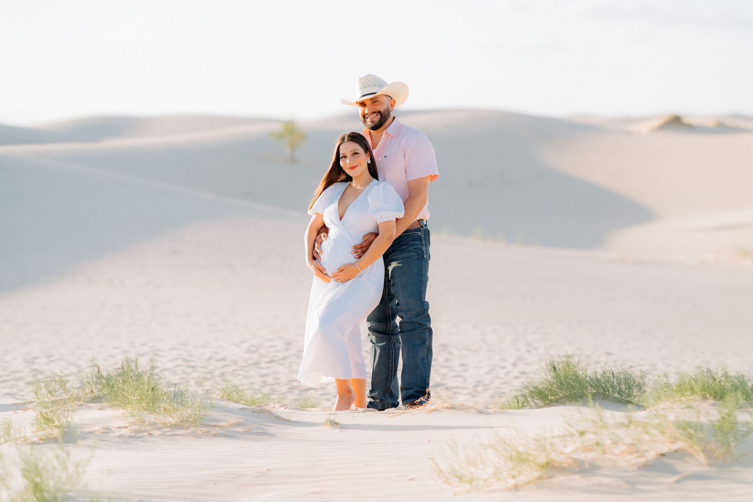 Expecting couple standing together on desert sand dunes in matching white and pink outfits.