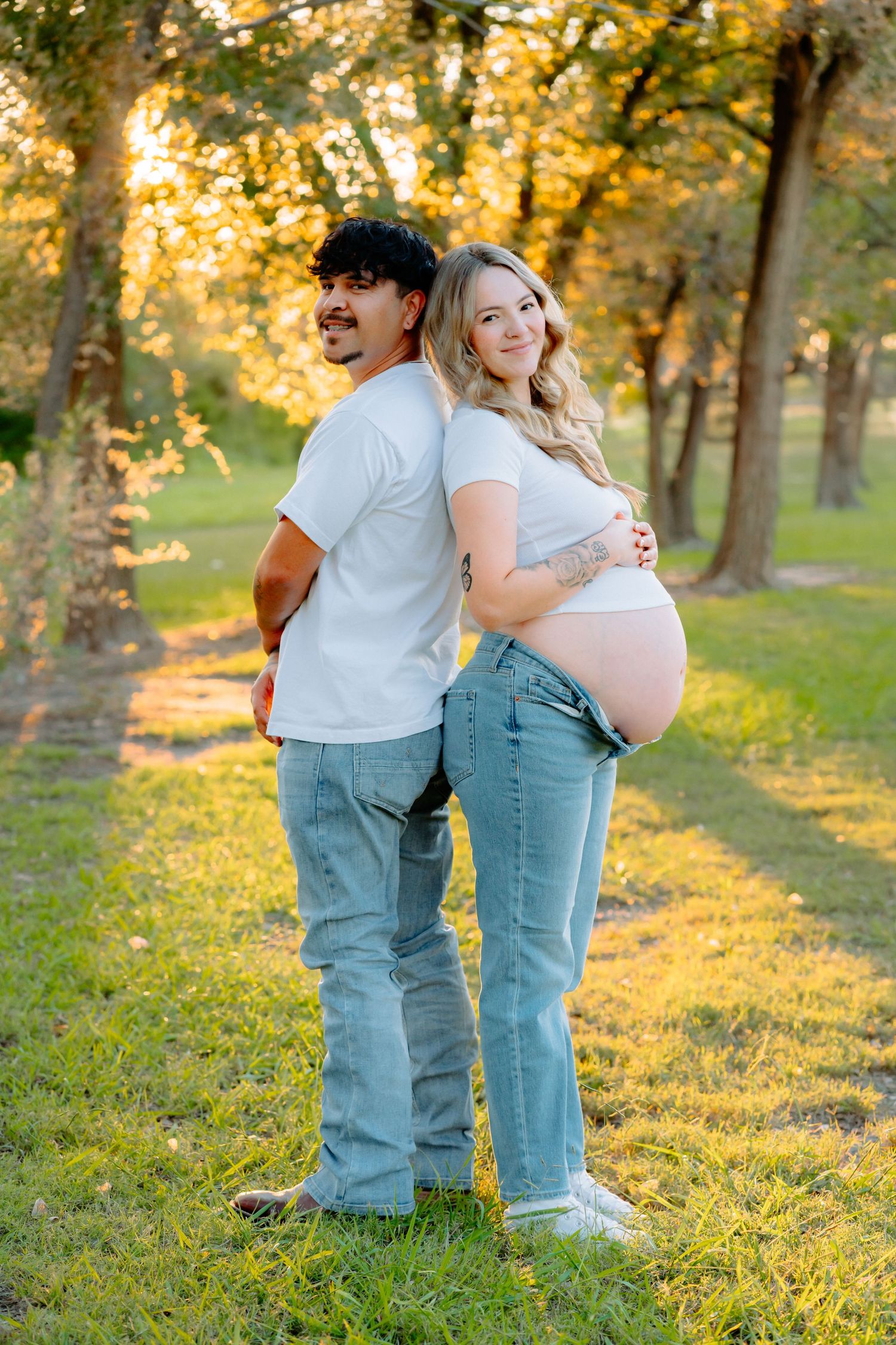 Couple wearing matching white shirts and jeans embrace in a sunlit autumn park setting during golden hour.