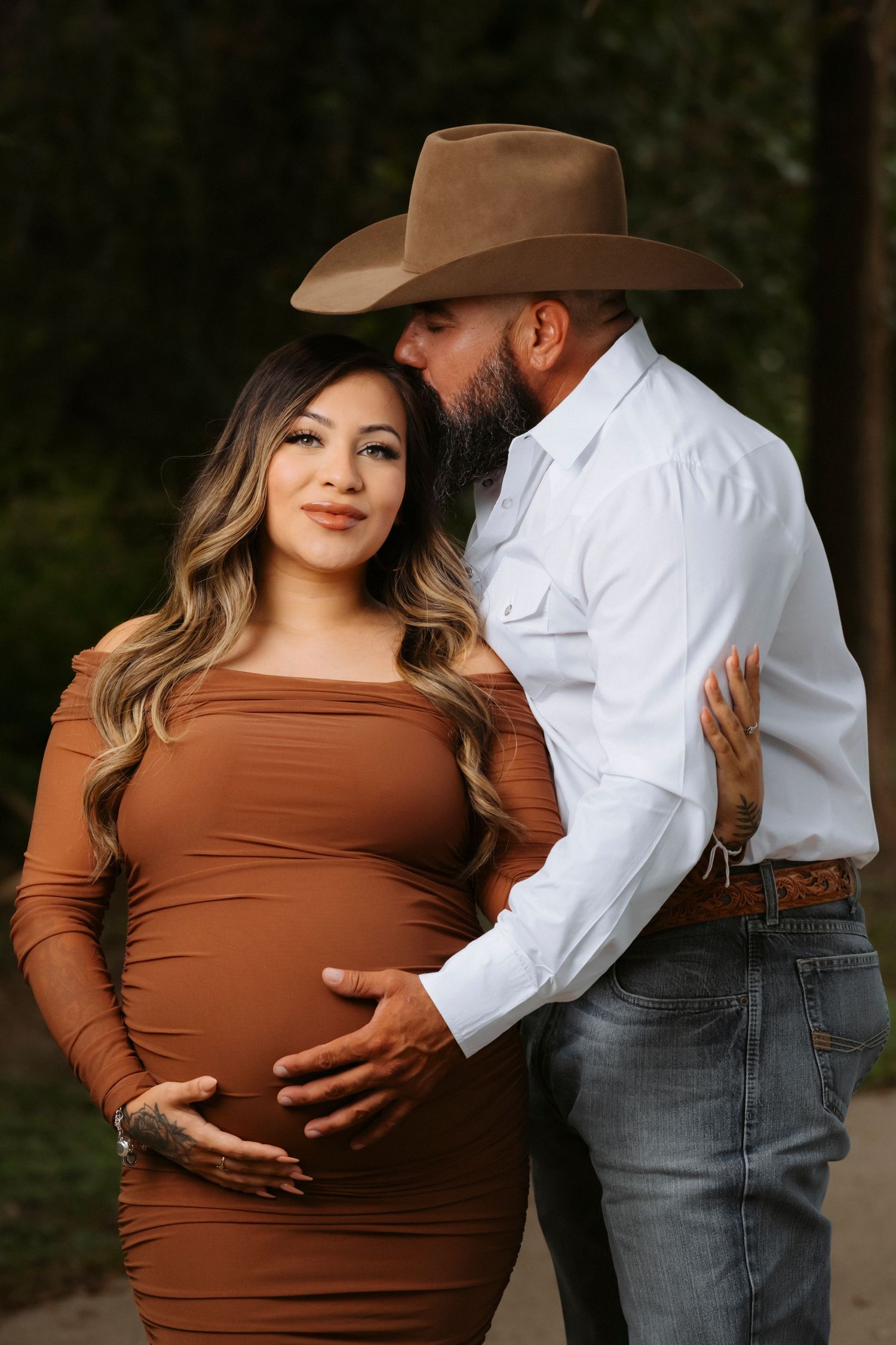 Expectant couple in western attire embraces during outdoor maternity photoshoot in brown dress and white shirt.