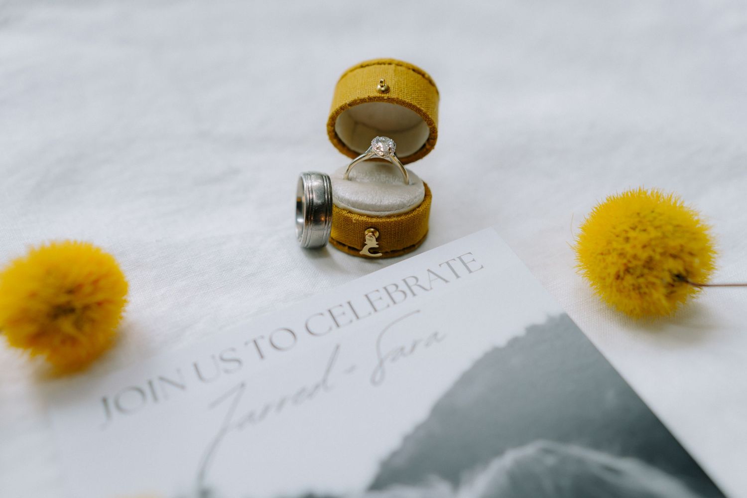Wedding rings in yellow ring box with pom pom decorations on invitation suite.