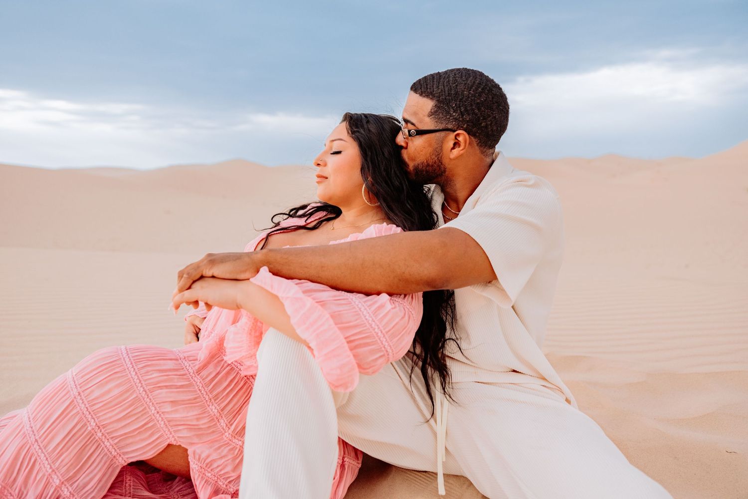 Romantic couple embraces while sitting on sand dunes in matching white and pink outfits during desert photoshoot.