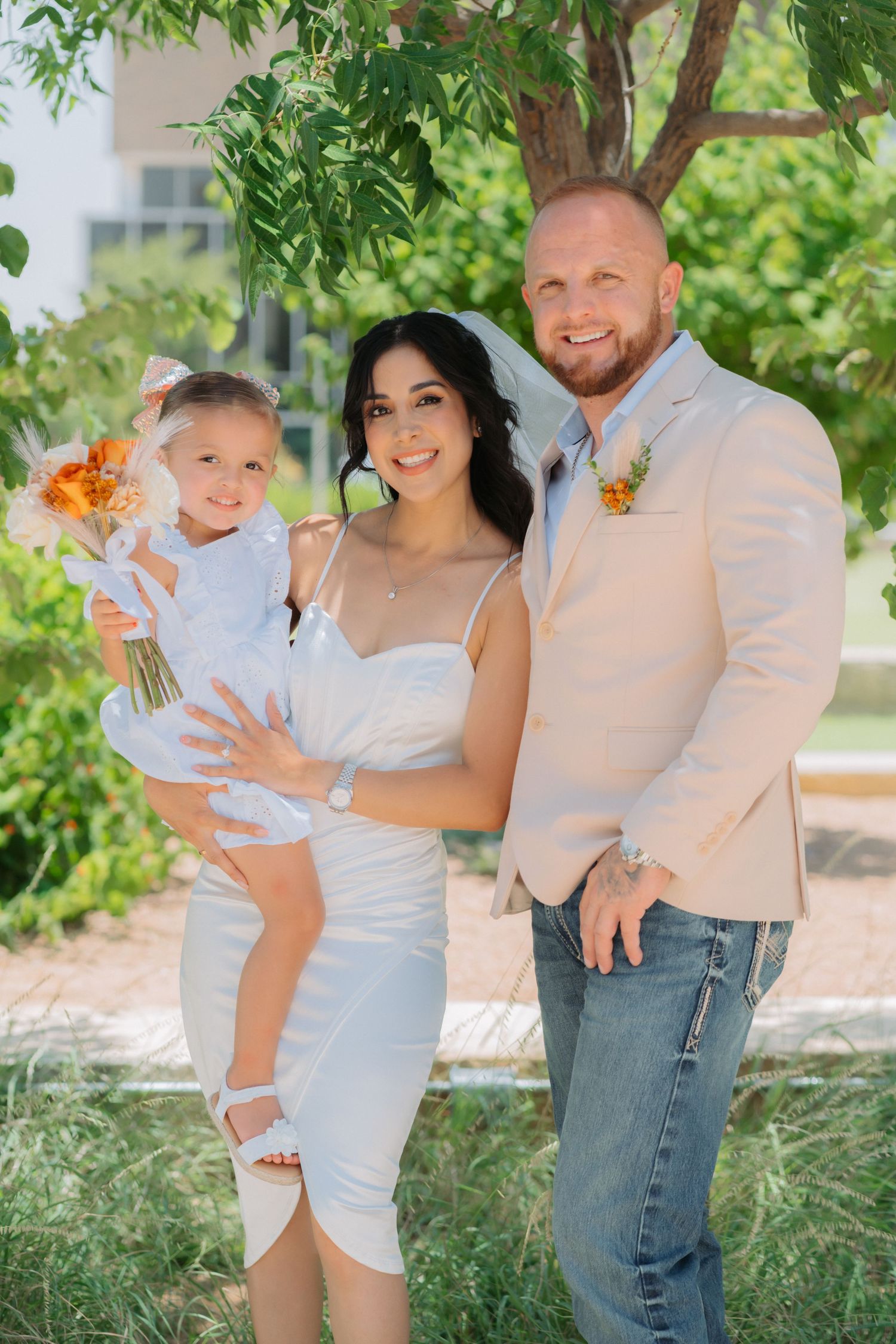 A beautiful outdoor family portrait under a green tree with orange floral accents and light colored attire.