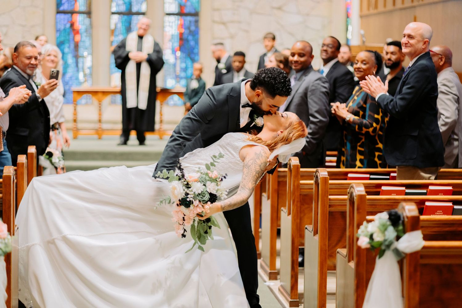 A couple shares a romantic dip kiss in the aisle of a church during their wedding ceremony as guests look on.