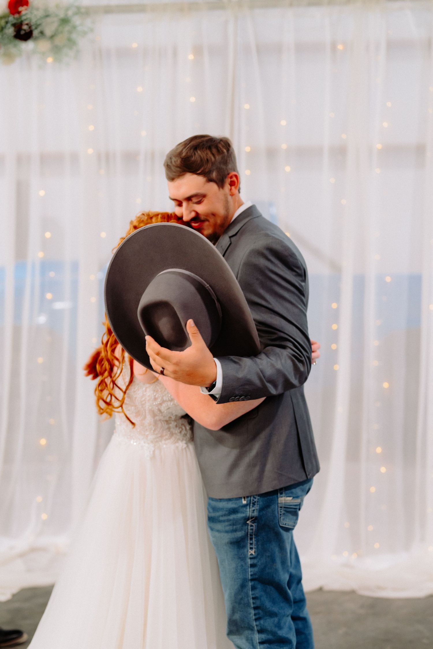 Intimate wedding dance with cowboy hats against white curtain backdrop with twinkle lights.