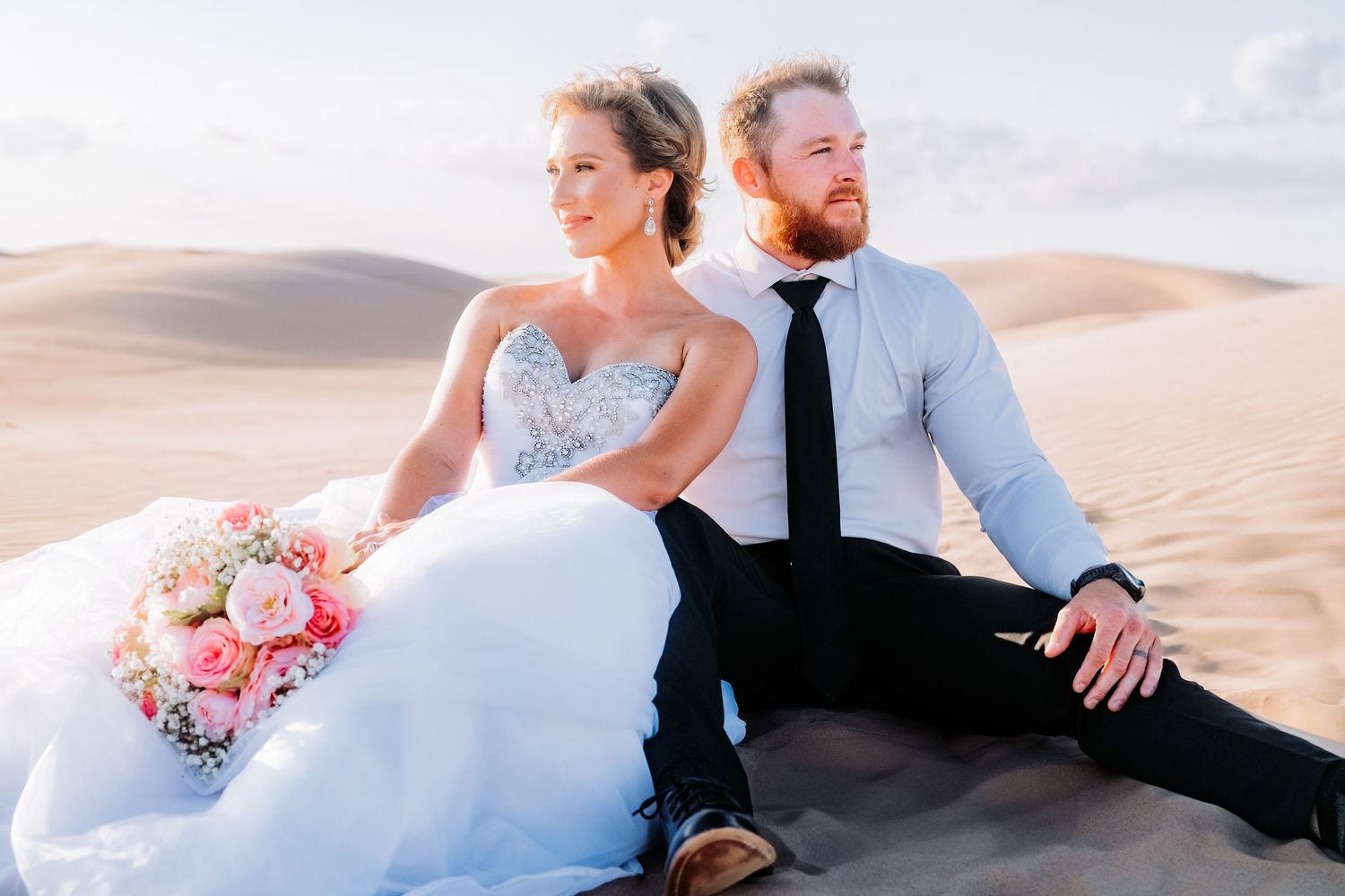 Newlyweds sitting together on sand dune during desert wedding photoshoot.