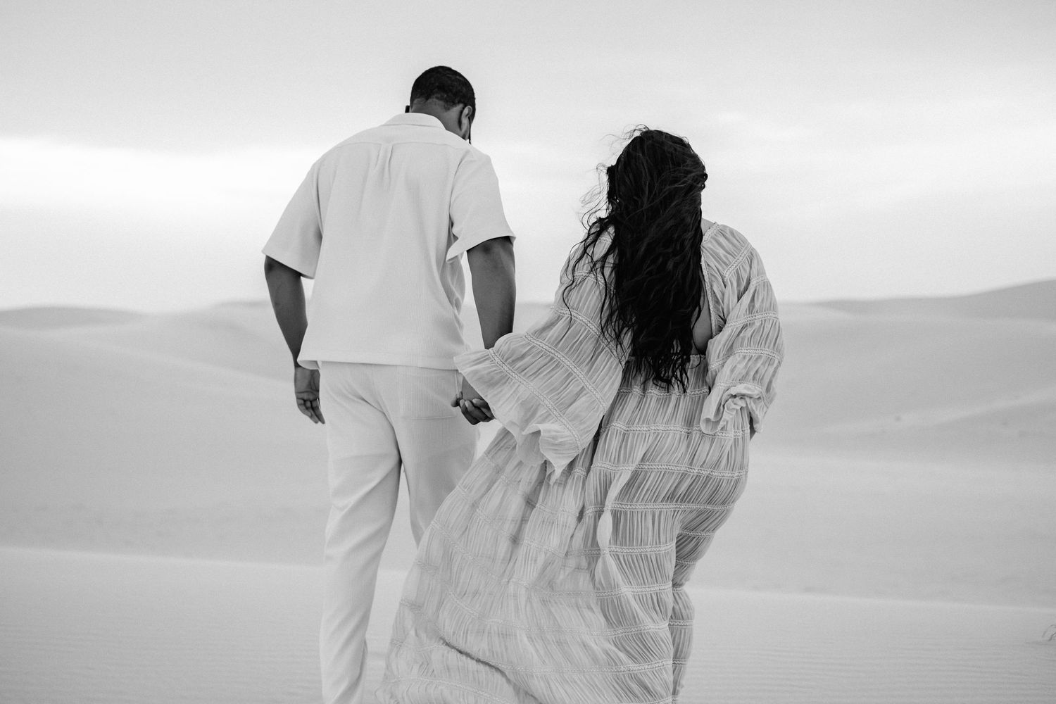 Black and white photo of couple holding hands while walking through desert sand dunes.