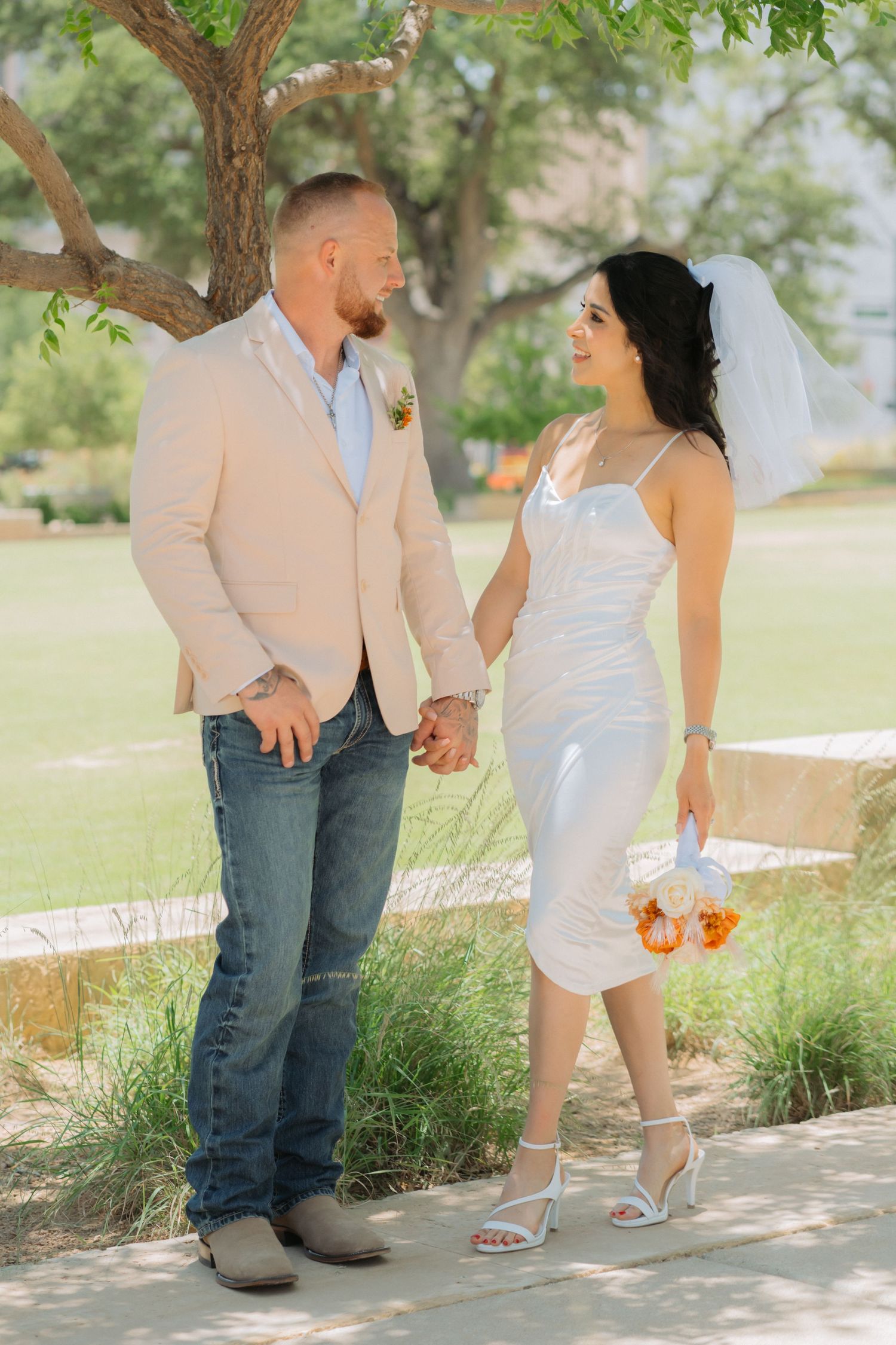 A couple in casual wedding attire stands together under a tree holding hands and smiling at each other outdoors.