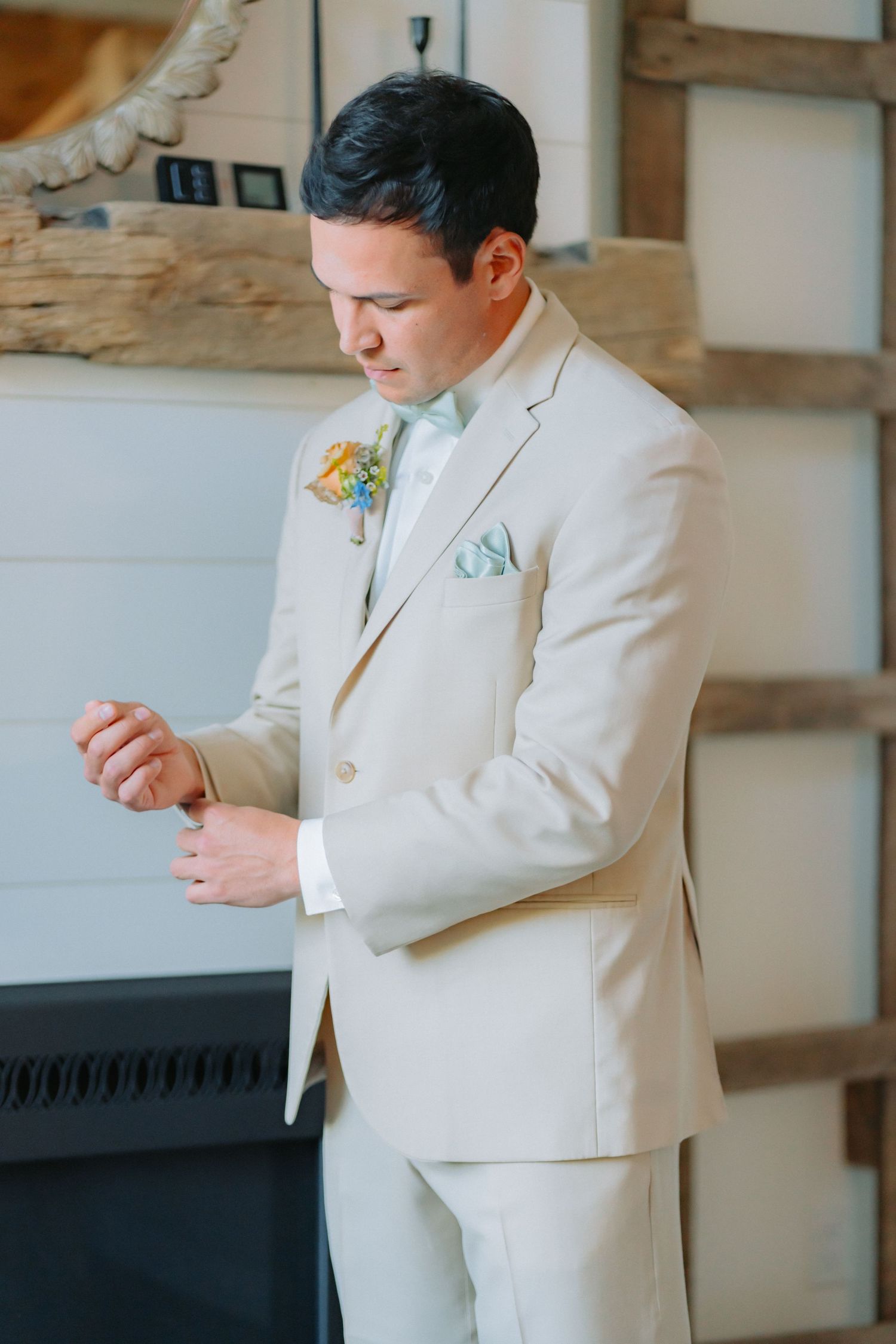 Groom in light beige suit adjusting cufflinks near fireplace.