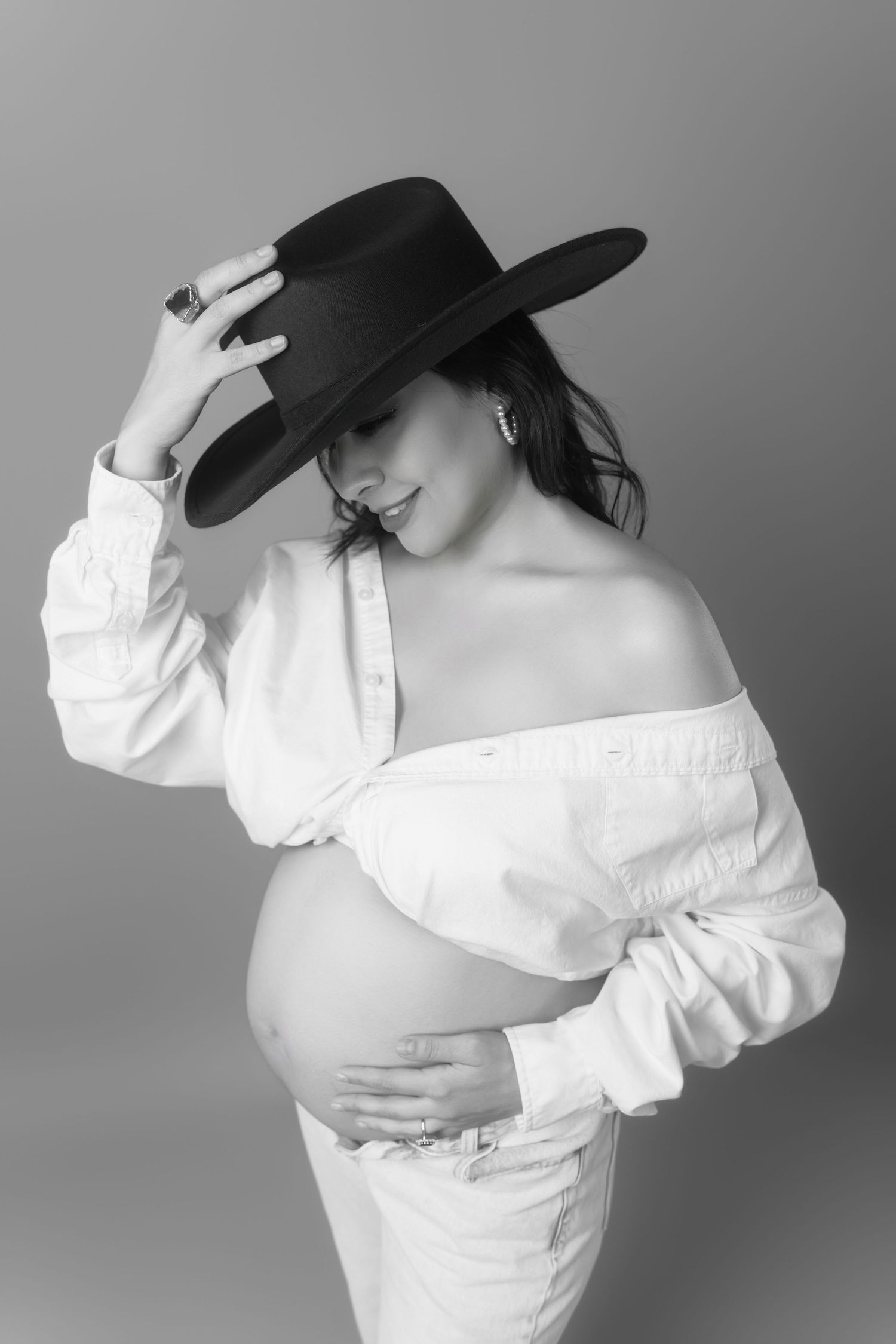 Black and white maternity photo featuring an elegant off-shoulder white dress and wide-brimmed black hat.