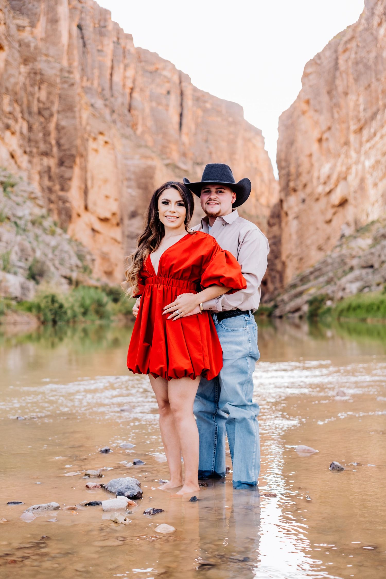 Couple standing in shallow water with canyon backdrop, woman in flowing red dress.