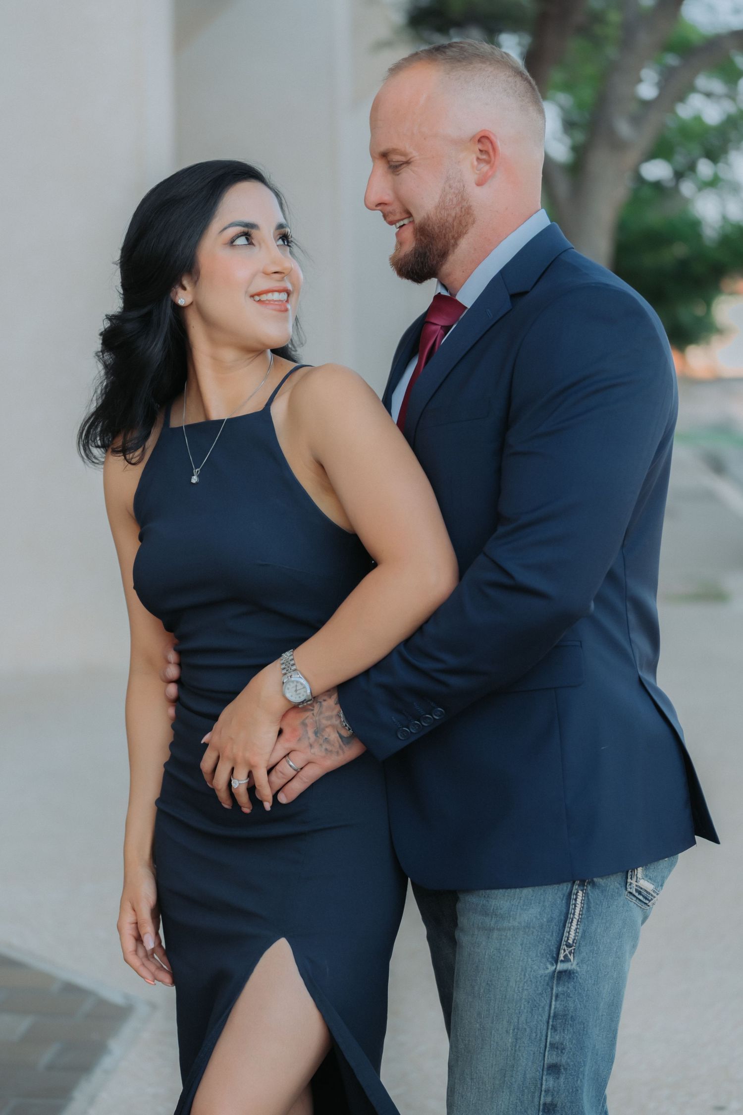 A couple poses together outdoors, with the woman wearing a black dress and the man in a navy blazer and red tie.