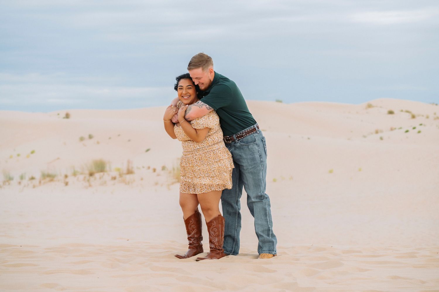 Couple embraces while standing together on sandy dunes during sunset photoshoot at the beach.