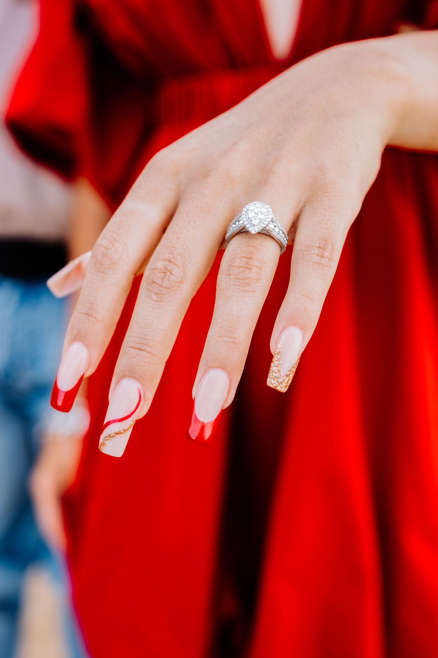 Close up of an elegant diamond engagement ring on a feminine hand with french manicured nails against red fabric.