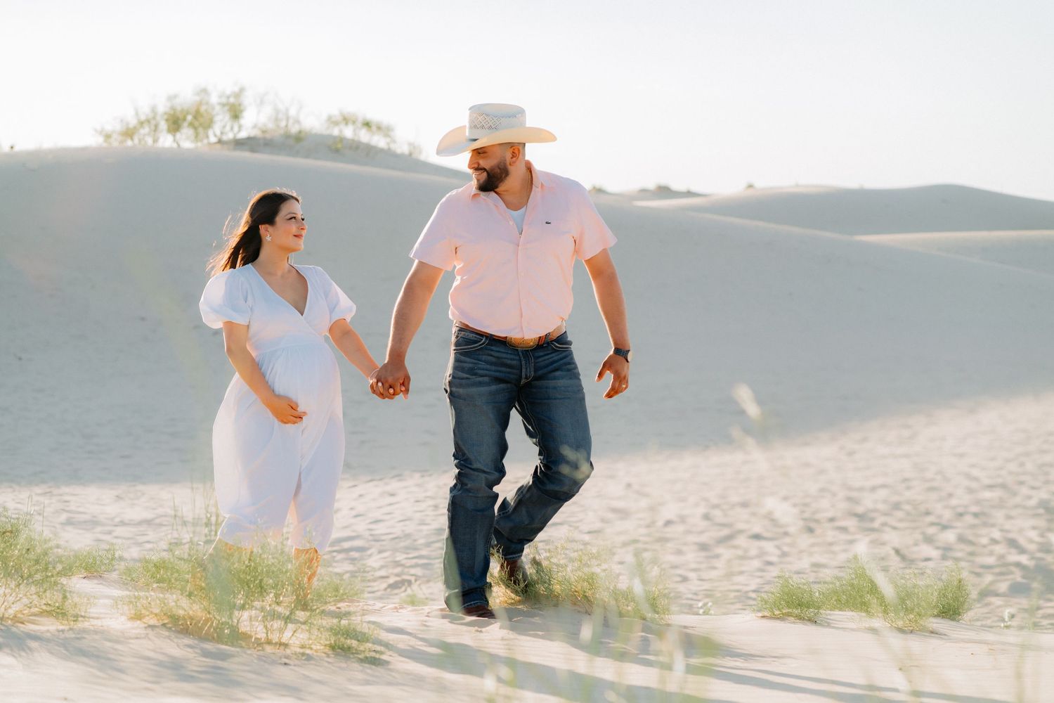 Couple walking hand in hand across desert sand dunes during maternity session.