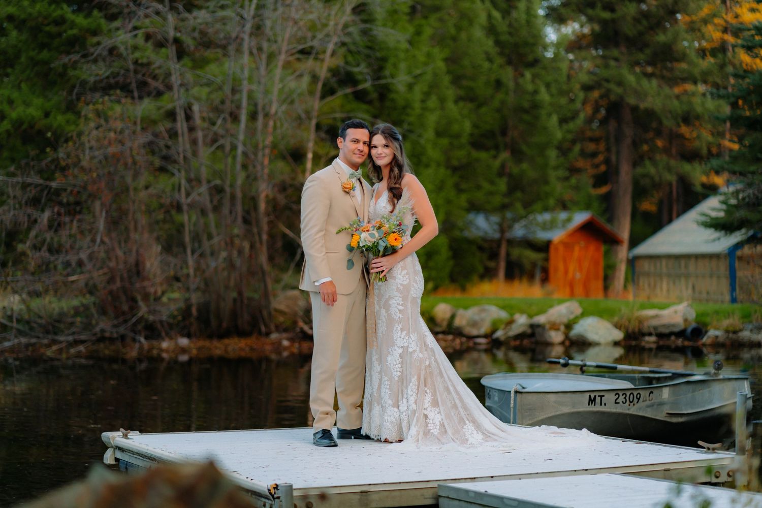Wedding couple stands together on a wooden dock overlooking a serene lake surrounded by autumn trees.