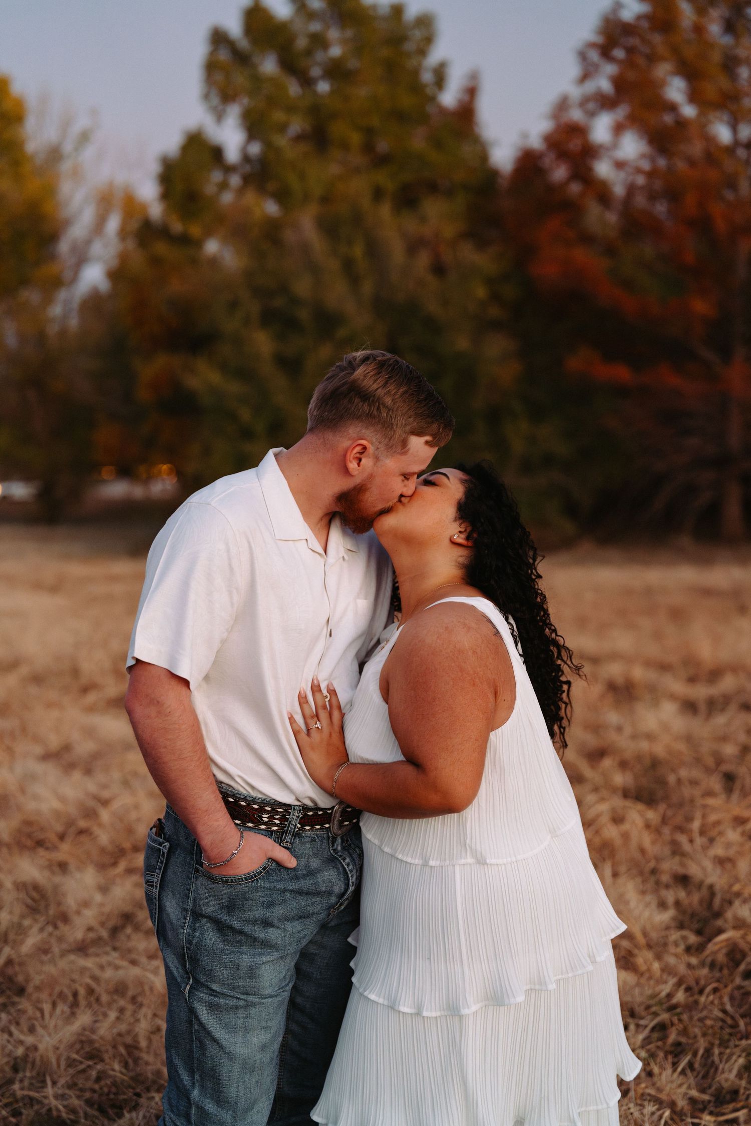 A romantic couple embraces in a field during autumn with warm golden light and fall foliage in the background.