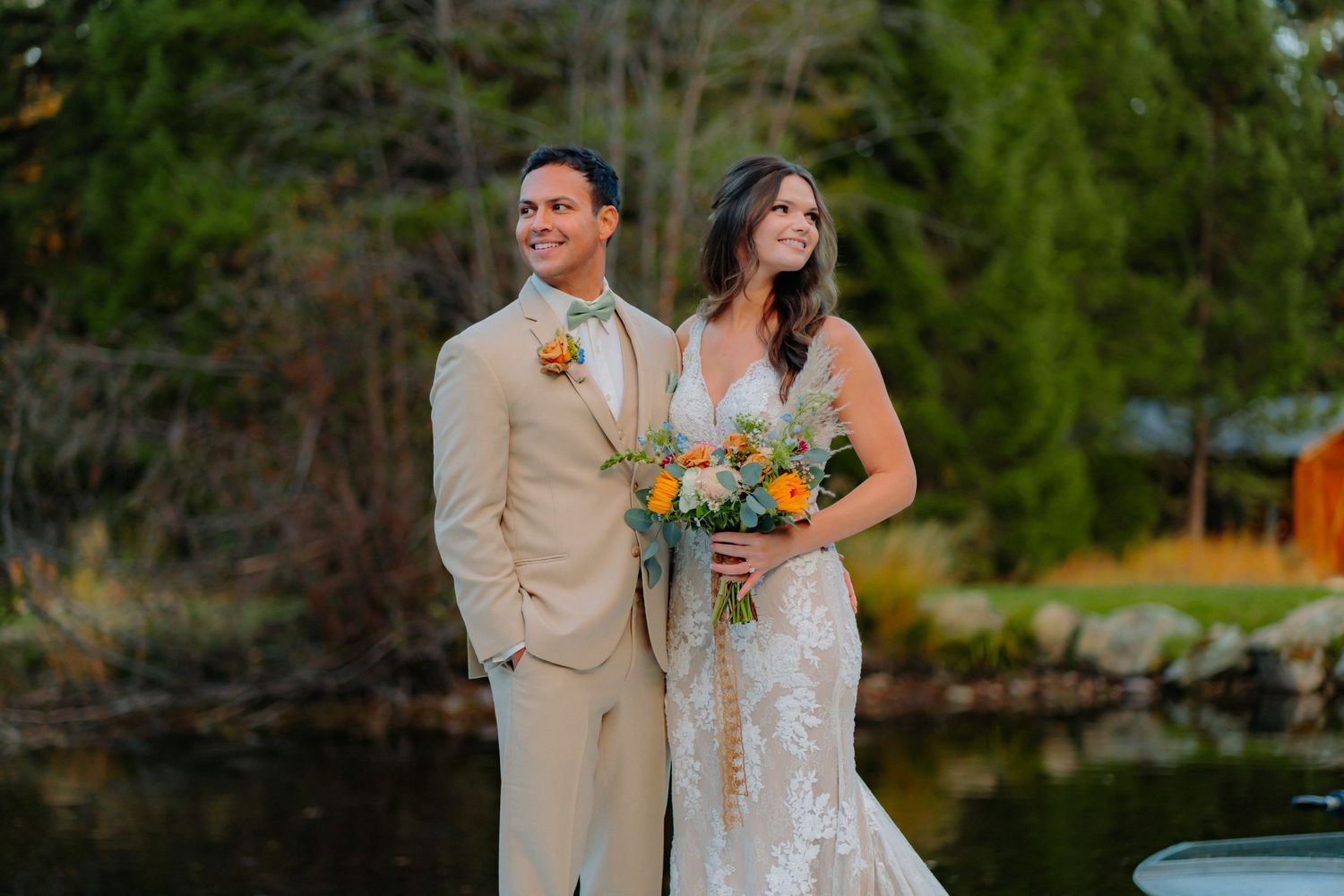 Wedding couple in tan suit and lace dress pose together on dock with autumn foliage in background.