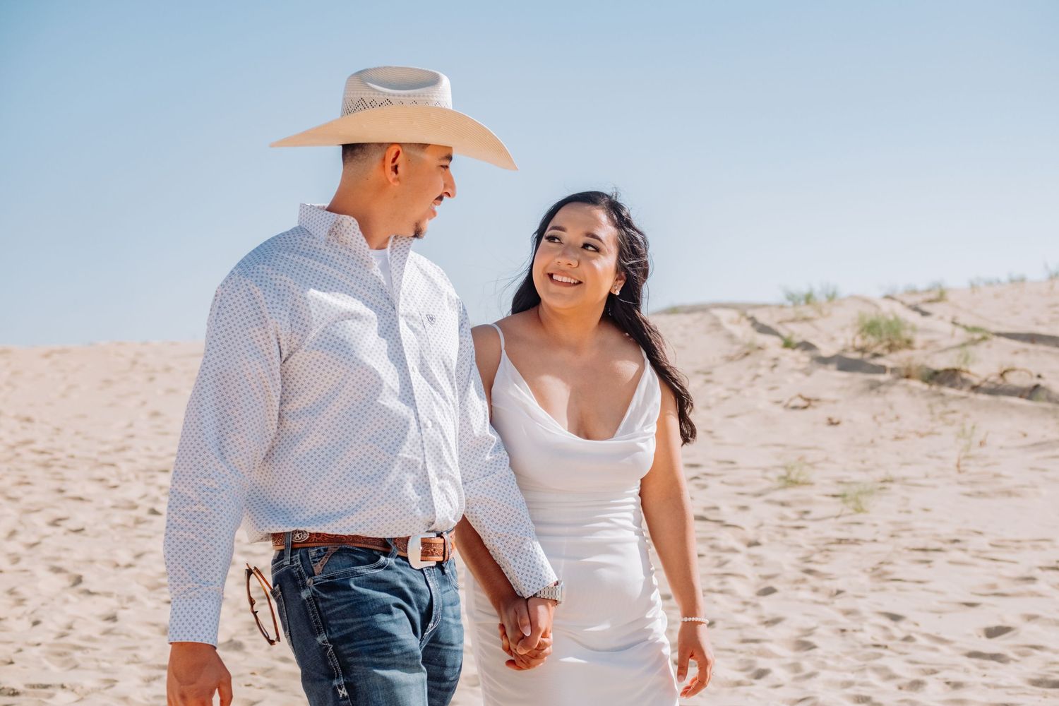 Couple walks hand in hand across desert sand dunes during photo session.