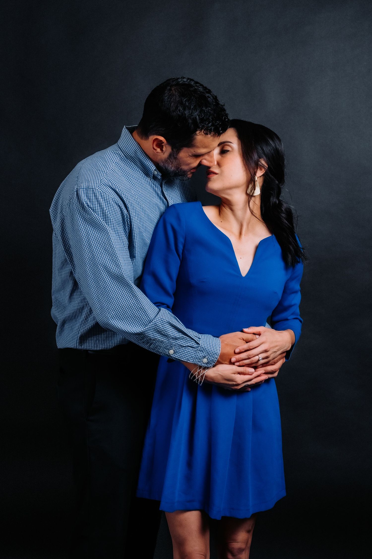 Romantic couple in blue dress and denim shirt embrace intimately against dark background.