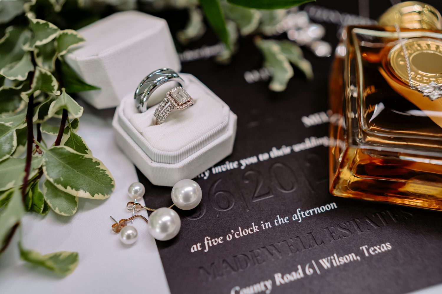 Wedding invitation suite with elegant calligraphy, rings, perfume bottle and greenery on dark surface.