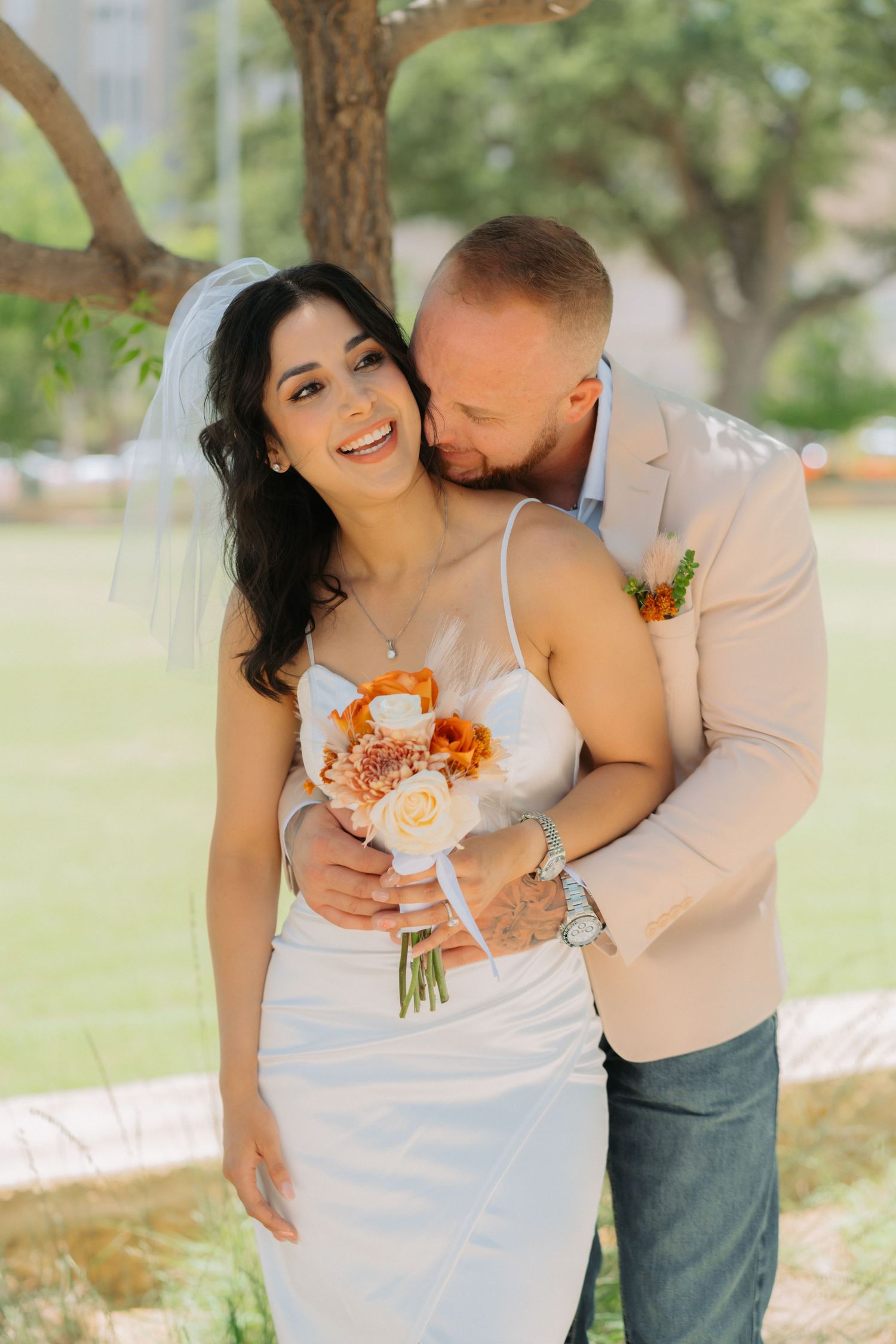 Joyful couple embraces outdoors during wedding photoshoot with bride holding peach and white floral bouquet.