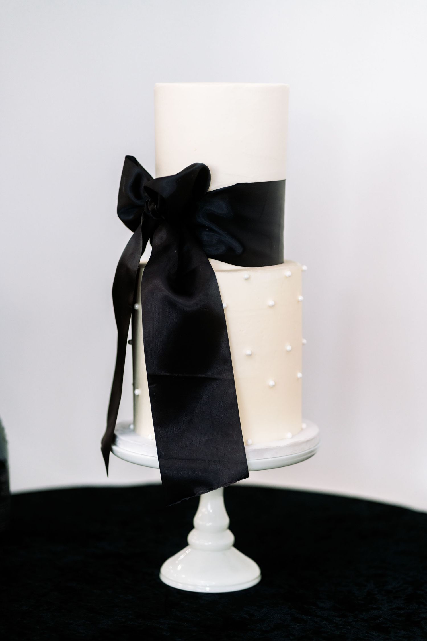 Elegant white wedding cake with black satin bow on white pedestal against dark background.