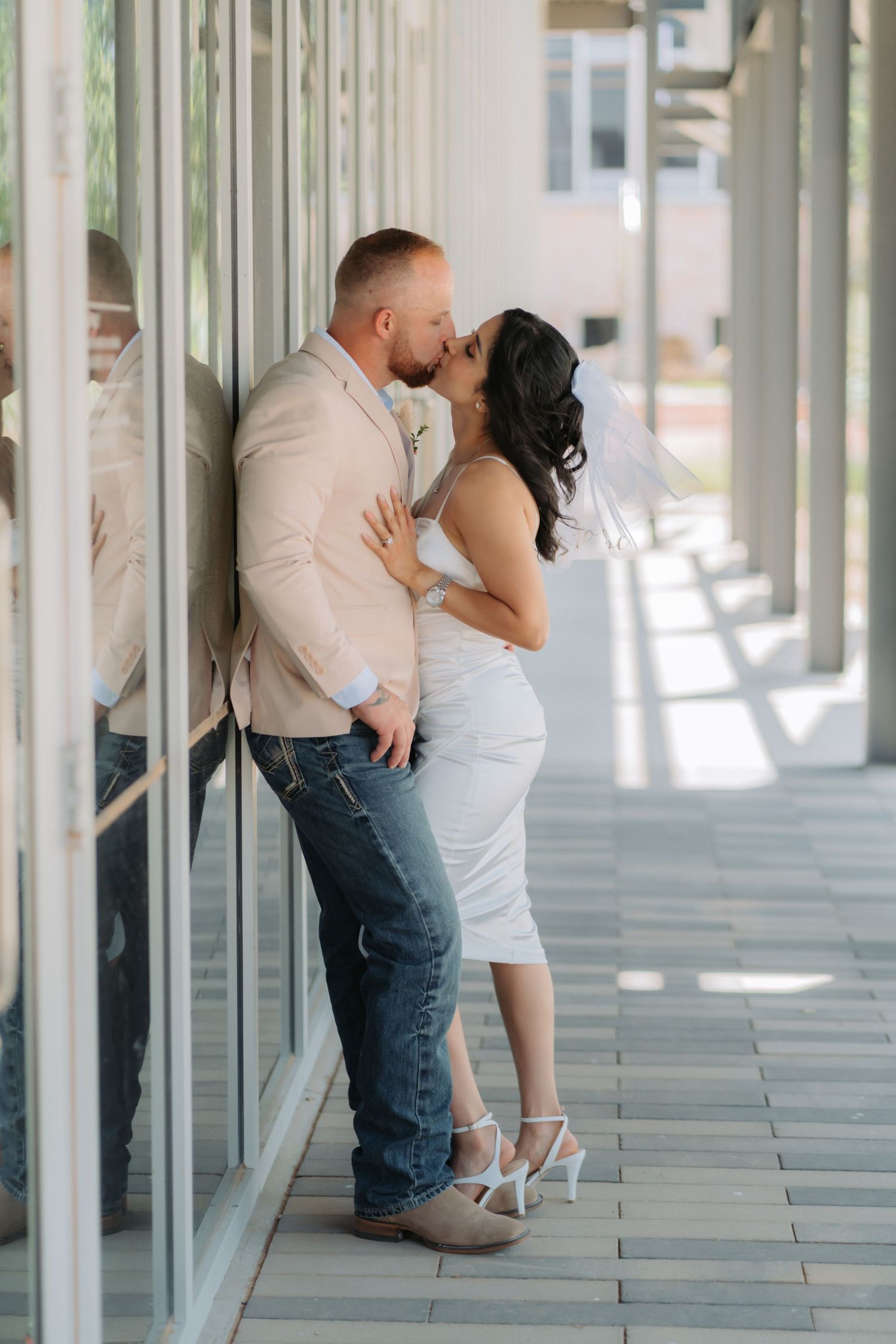 A romantic moment captured between a couple in formal wear standing in a sunlit hallway with glass windows.