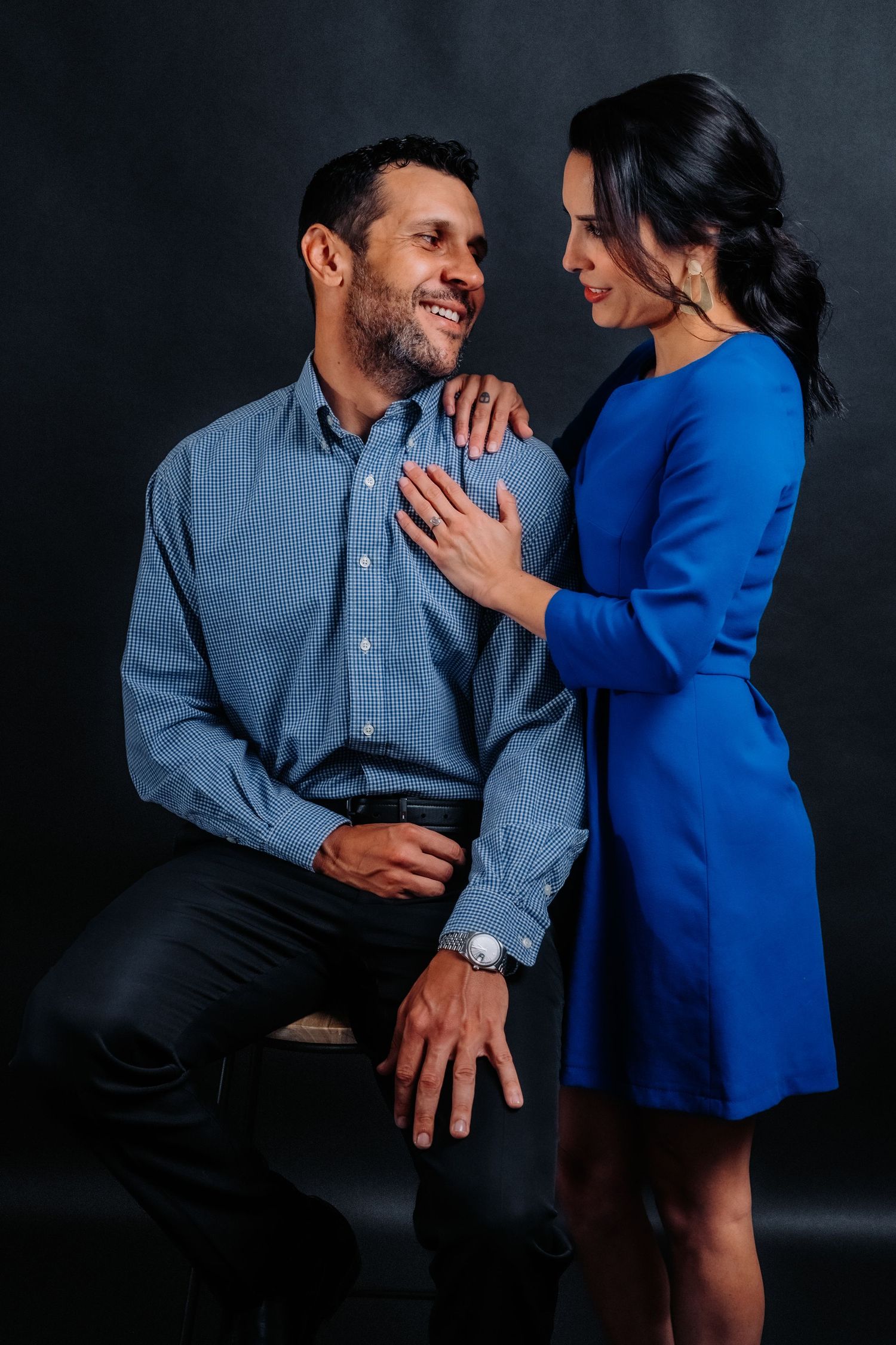 Stylish couple poses together, with woman in blue dress and man seated wearing chambray shirt.
