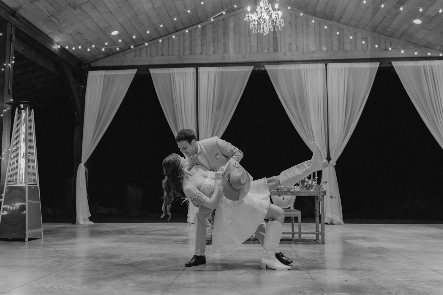Black and white photo of couple performing dramatic dip dance move at wedding reception under string lights.