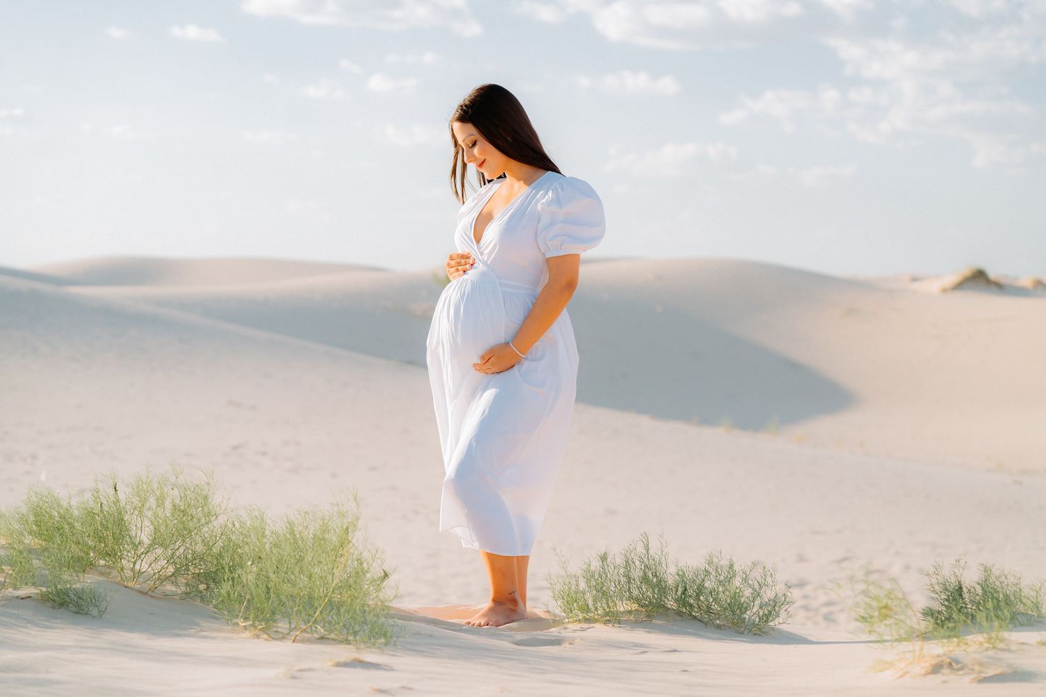 Pregnant woman in white dress standing alone on sand dunes during sunset photoshoot.