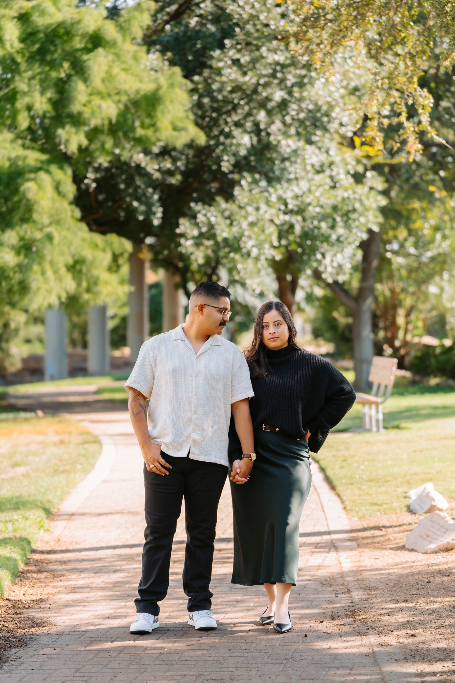 A couple takes a romantic stroll along a tree-lined path in a sunlit park.
