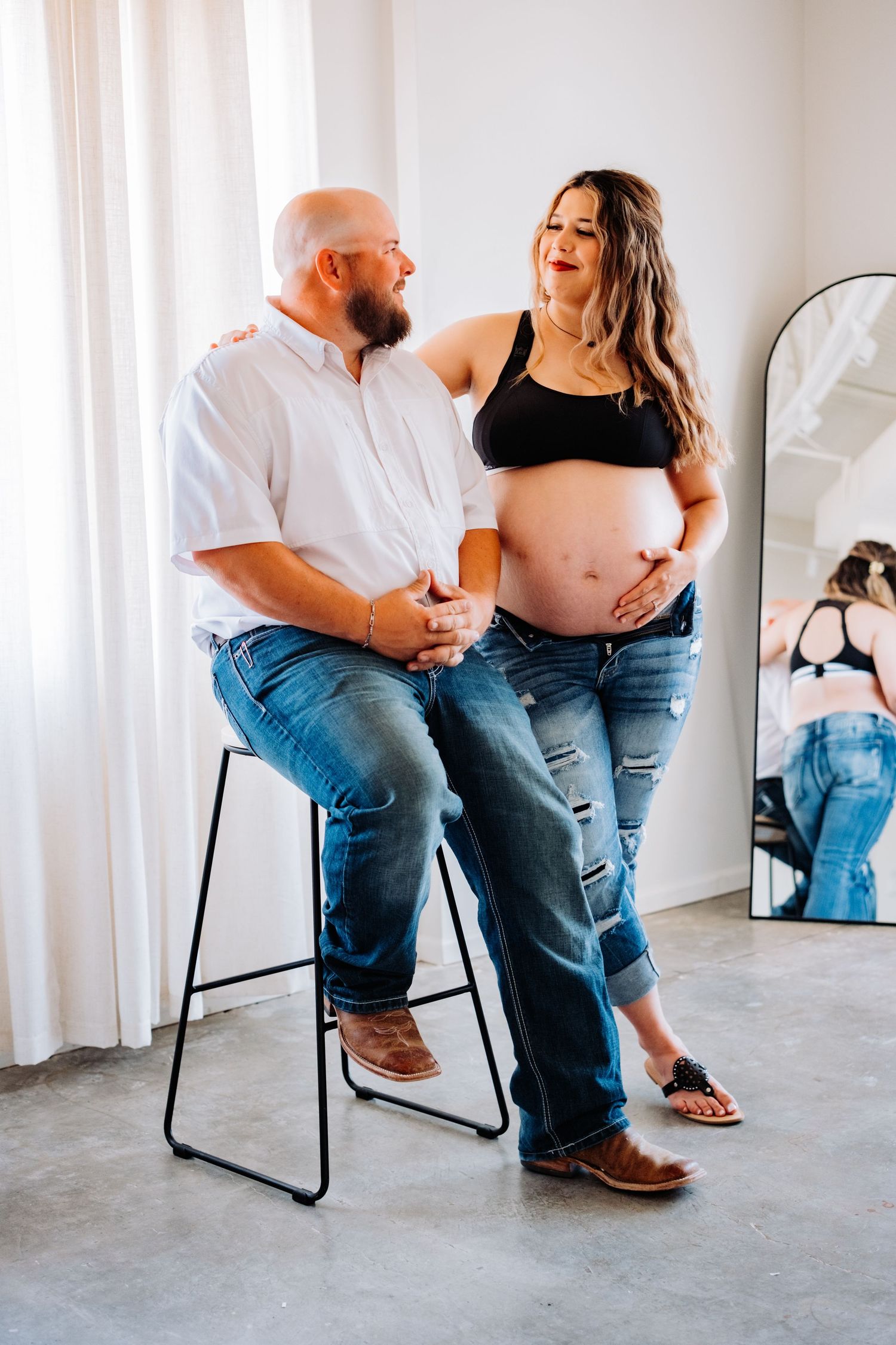 Casual maternity photo of couple sitting together on modern barstool in bright indoor setting.