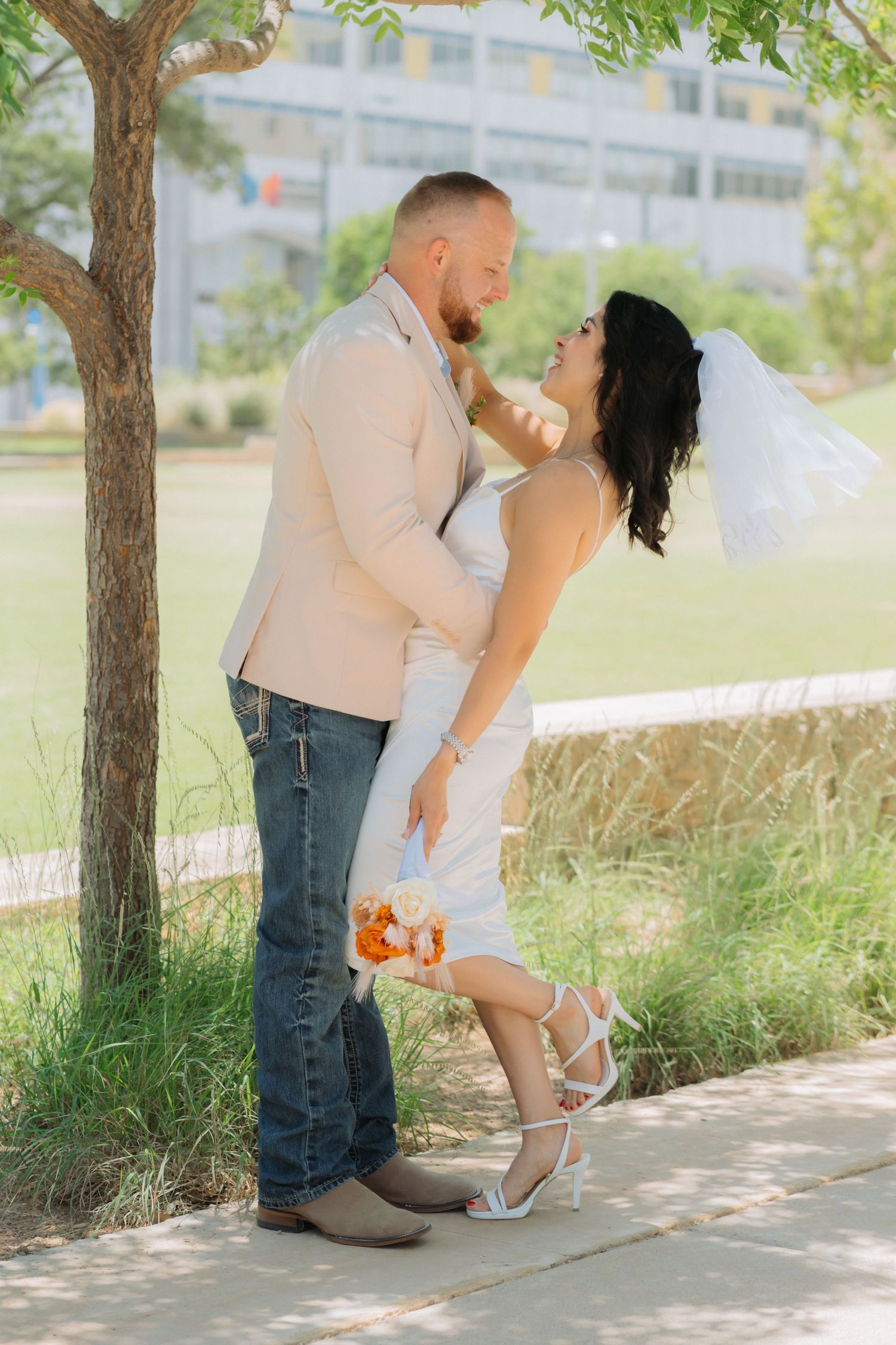 A couple shares a tender moment for their wedding beside a tree on a sunny day in a park setting at Centennial Park in Midland