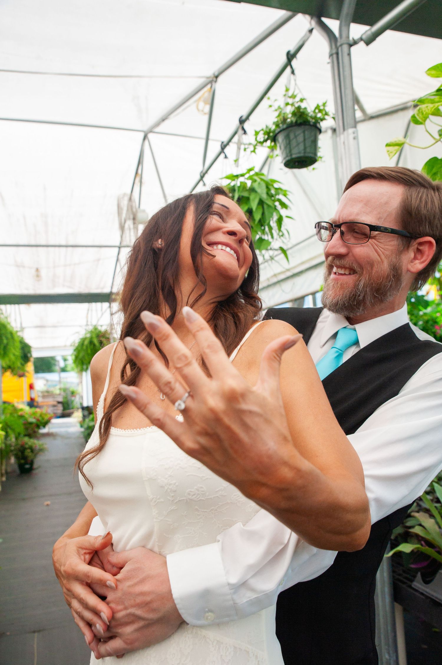 A joyful moment captured in a greenhouse as a couple laughs and embraces while showing off a ring.