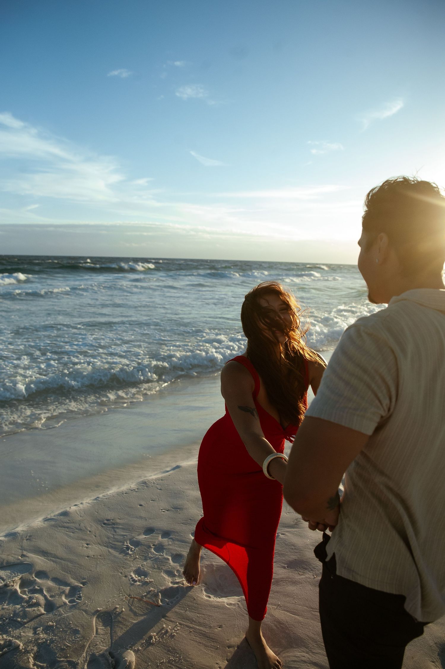 Two people walking along a sunny beach at sunset with waves crashing on the shore in the background.