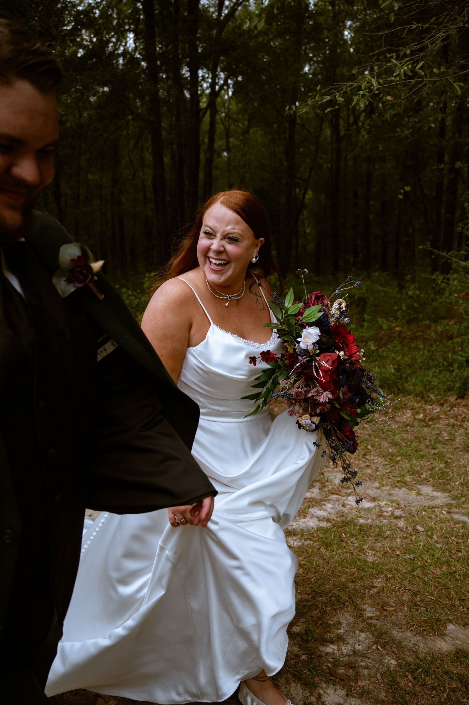 A bride in a white wedding dress laughs joyfully while holding a burgundy and pink bouquet in a wooded setting.