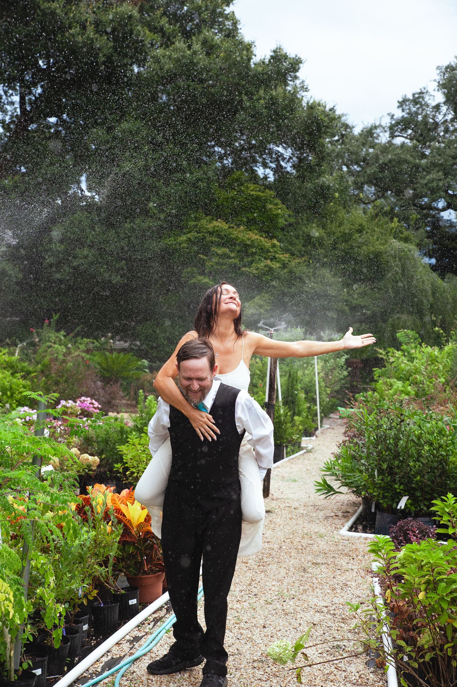 A couple shares a playful moment in a lush garden pathway with water sprinklers creating a magical mist effect.