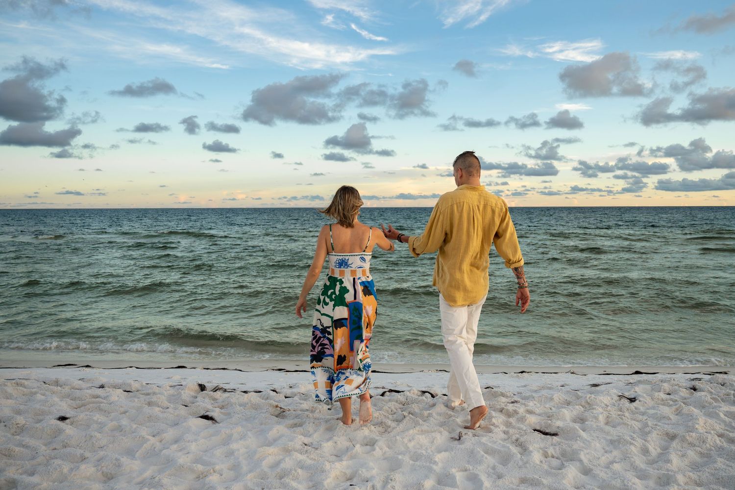Couple walking hand-in-hand on a white sandy beach at sunset, wearing colorful summer outfits near the ocean.