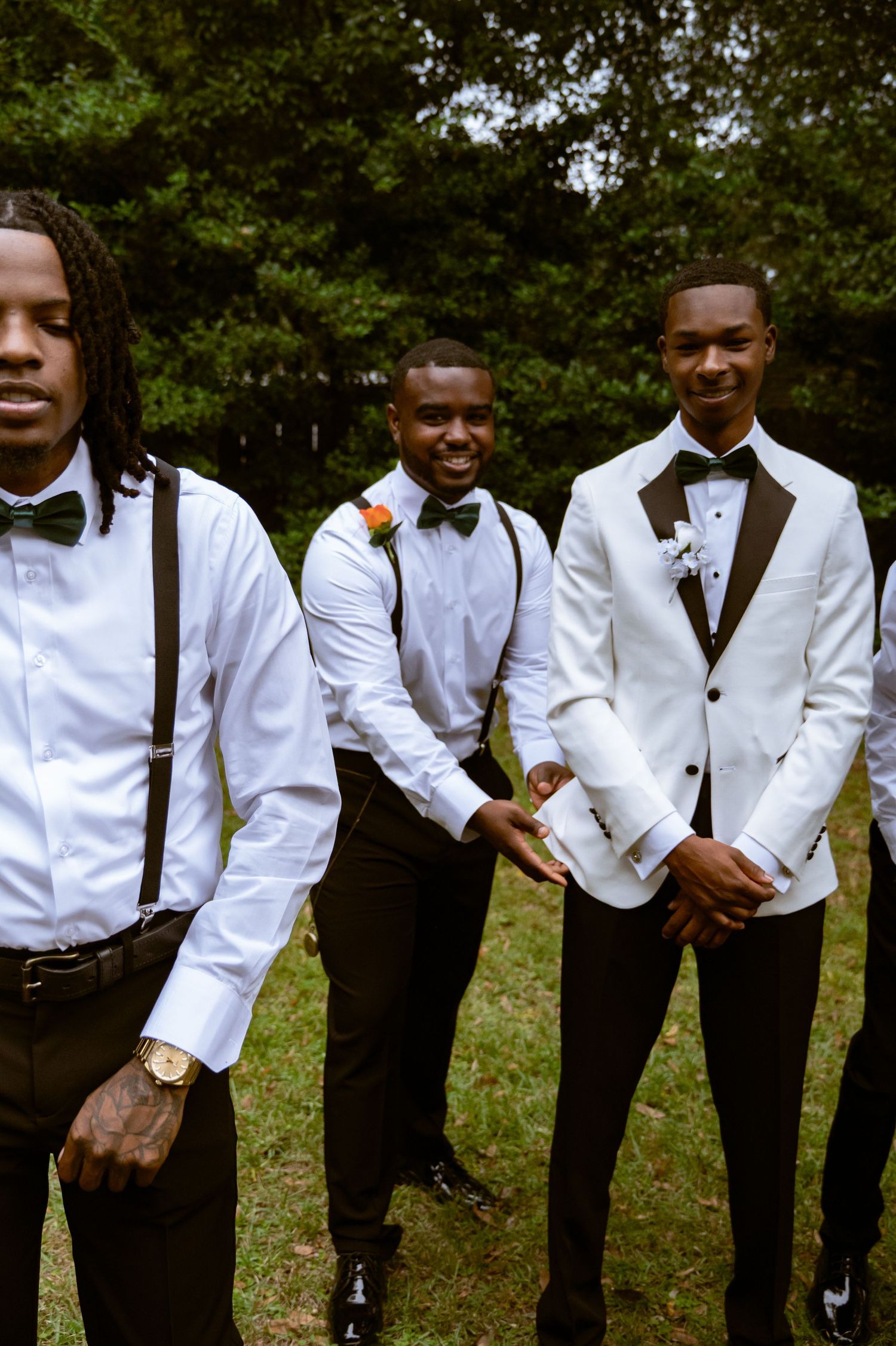 Three groomsmen in formal attire with white shirts, bow ties and suspenders pose outdoors against green foliage.