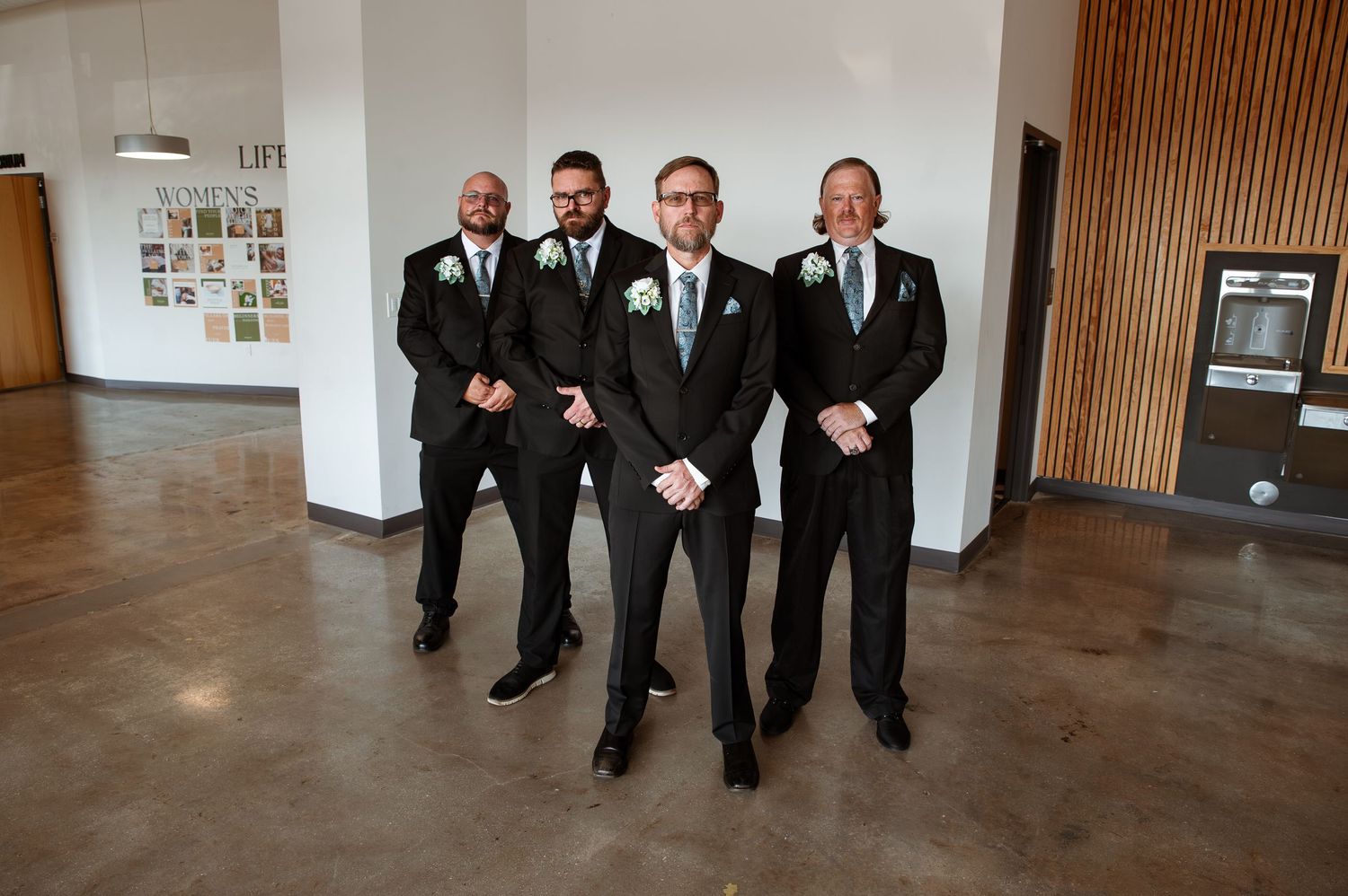 Groomsmen in black tuxedos pose together in a fun jumping photo at an indoor wedding venue.