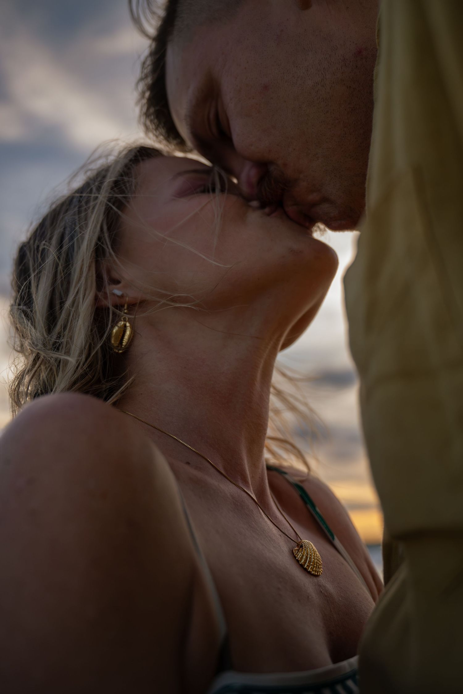 Couple sharing a romantic kiss at sunset, woman wearing gold earrings and necklace, warm golden light in background.