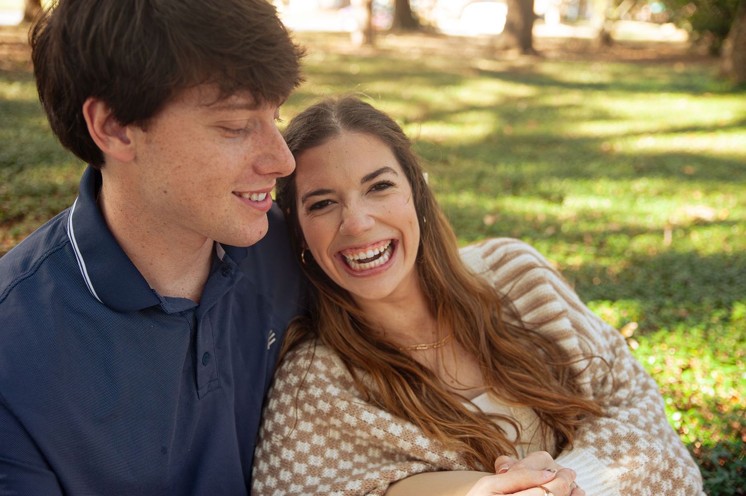 A young couple laughs together while sitting outdoors on a sunny day in a park setting.