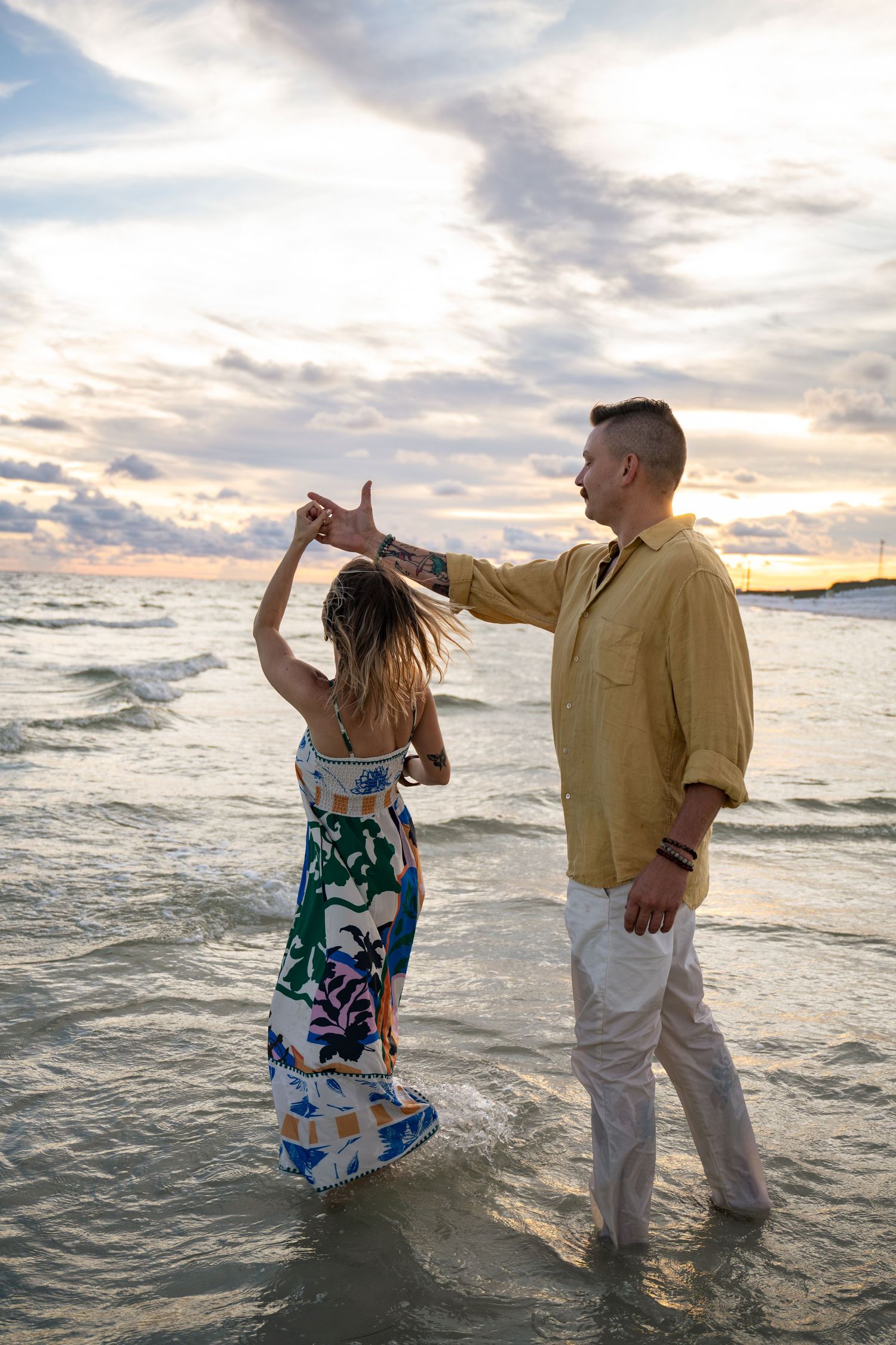 Couple dancing on beach at sunset, man in yellow shirt spinning woman in colorful floral dress, waves at their feet.